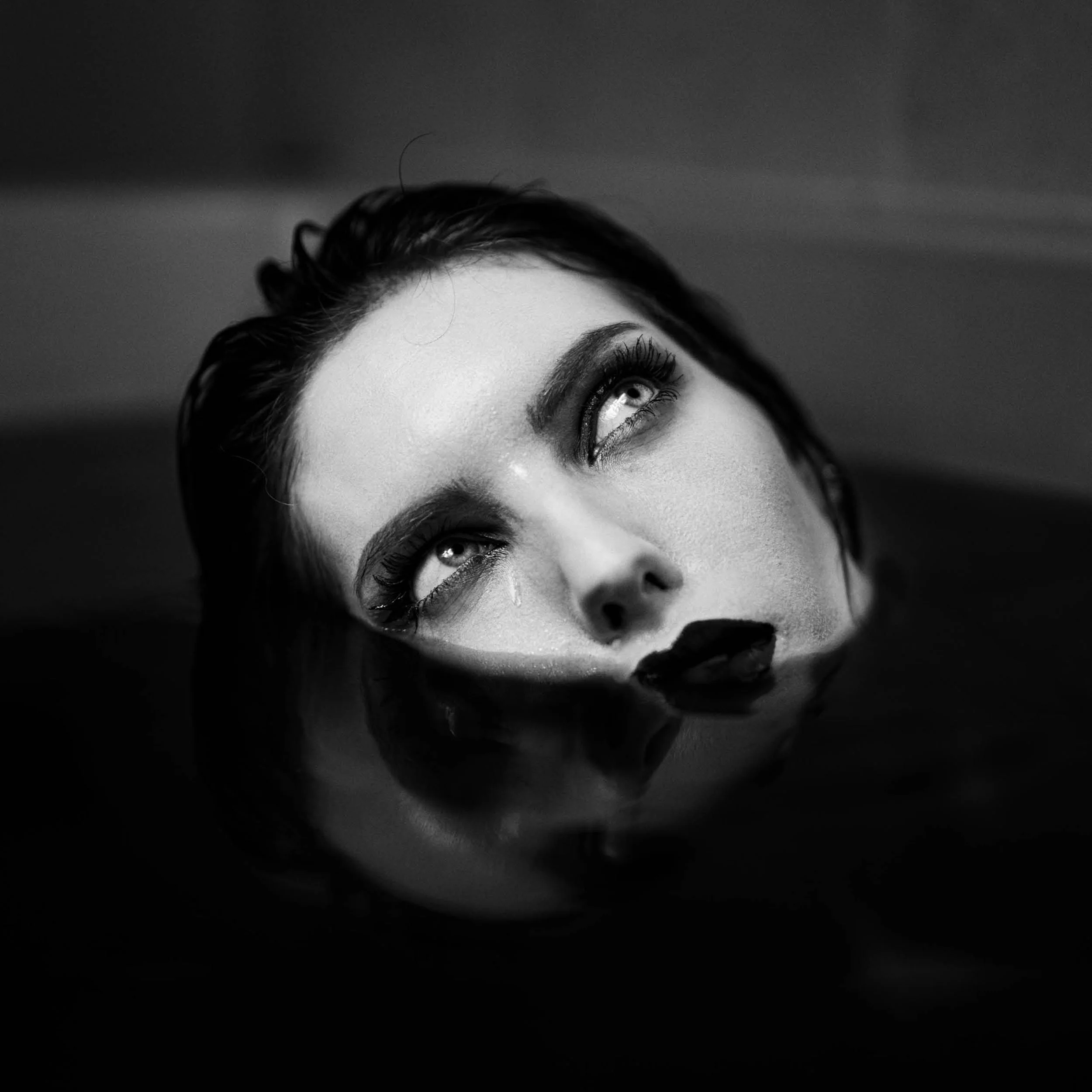 Black and white photo of a woman with makeup and tear rolling down her cheek, submerged in water with only her face visible above the surface.