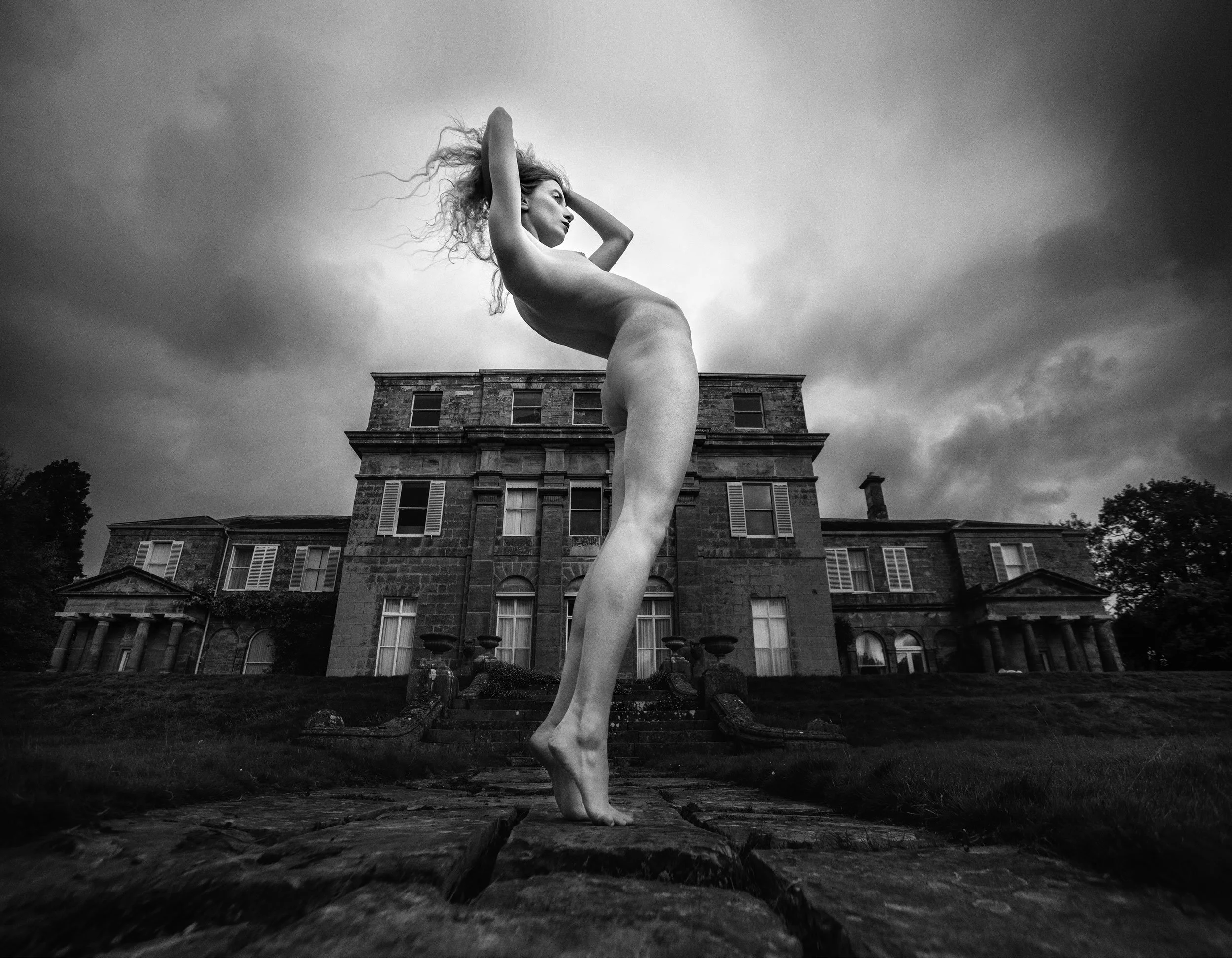 Black and white photo of a Lulu Lockhart standing outside Hammerwood Park Manor House in a dynamic nude pose 