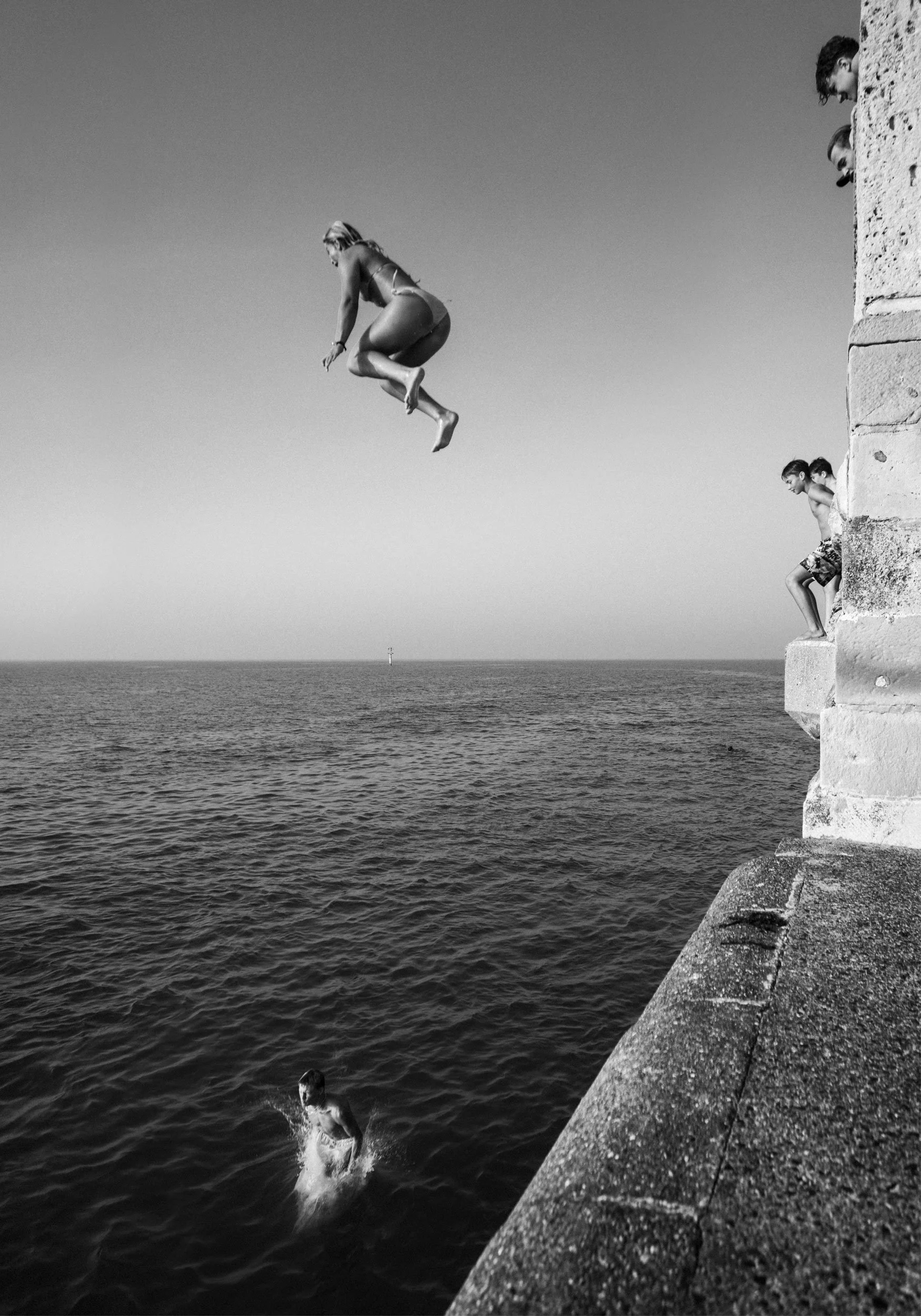 A black-and-white photo of people jumping from a stone pier into the water. One person is mid-air, and others are waiting to jump or already in the water. The scene is set by the sea with a buoy or marker in the distance.