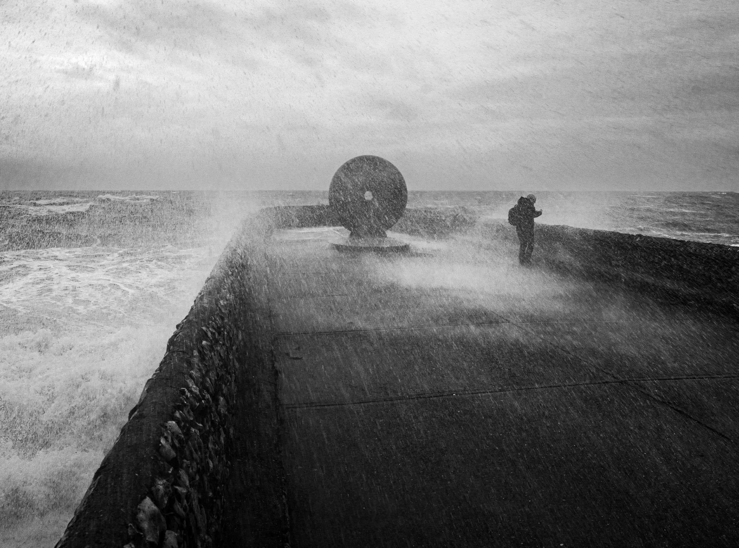 A person standing on a wind-swept seawall during a storm, with waves crashing and spray in the air.