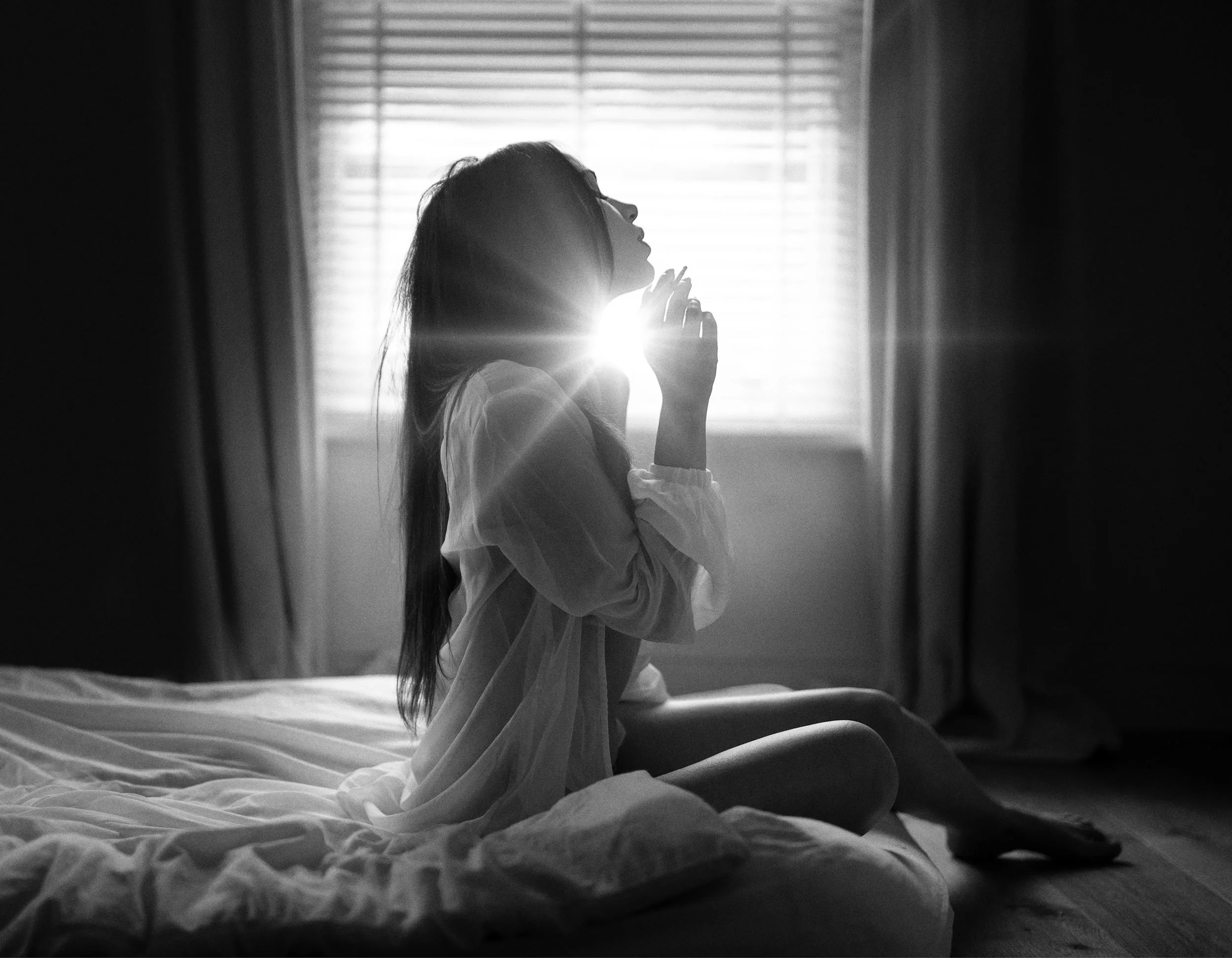 A woman sitting on a bed with her knees drawn up, touching her face in front of a window with blinds, backlit by sunlight.
