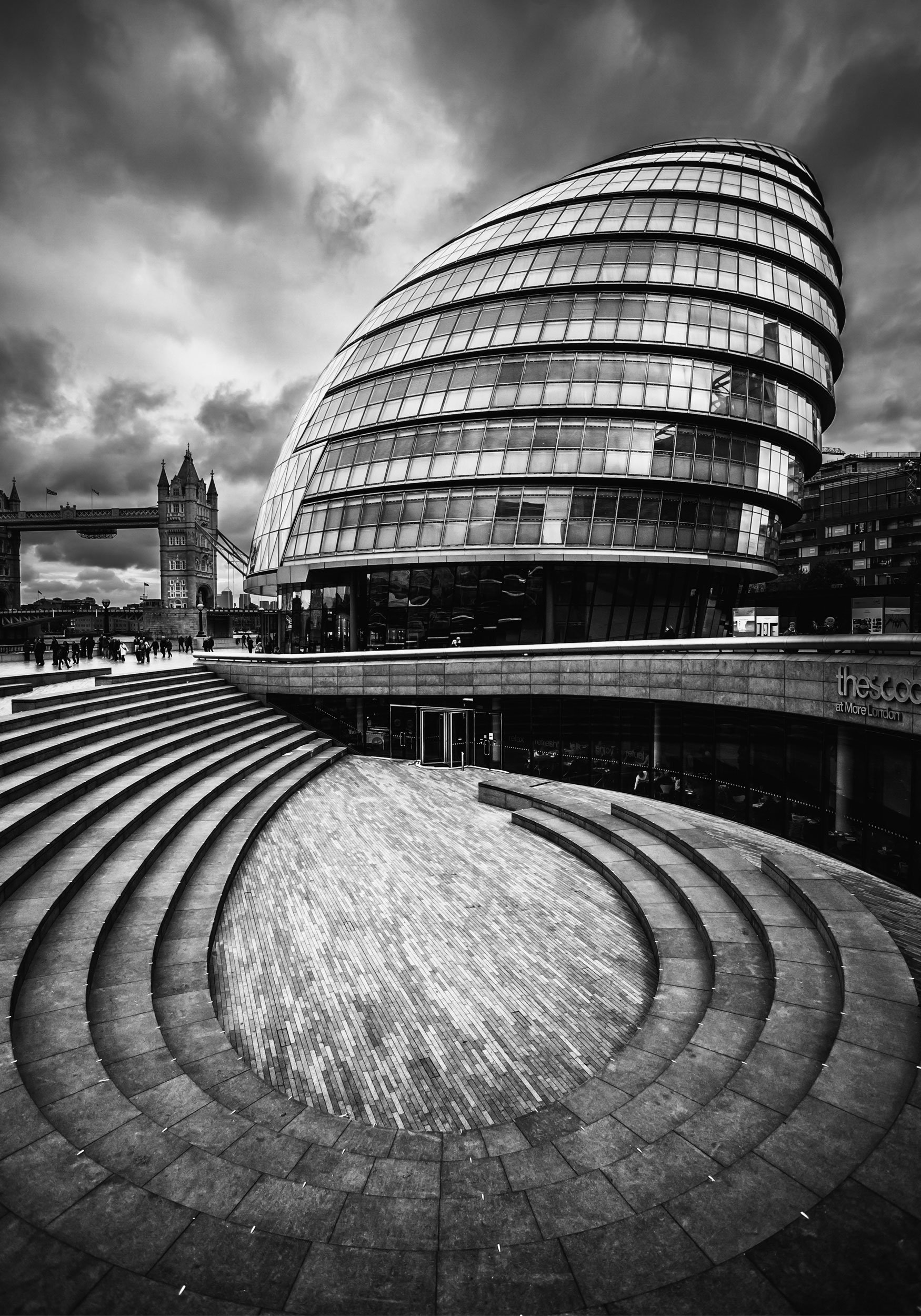 Black and white photo of City Hall in London with modern curved building in foreground and Tower Bridge in background under cloudy sky.