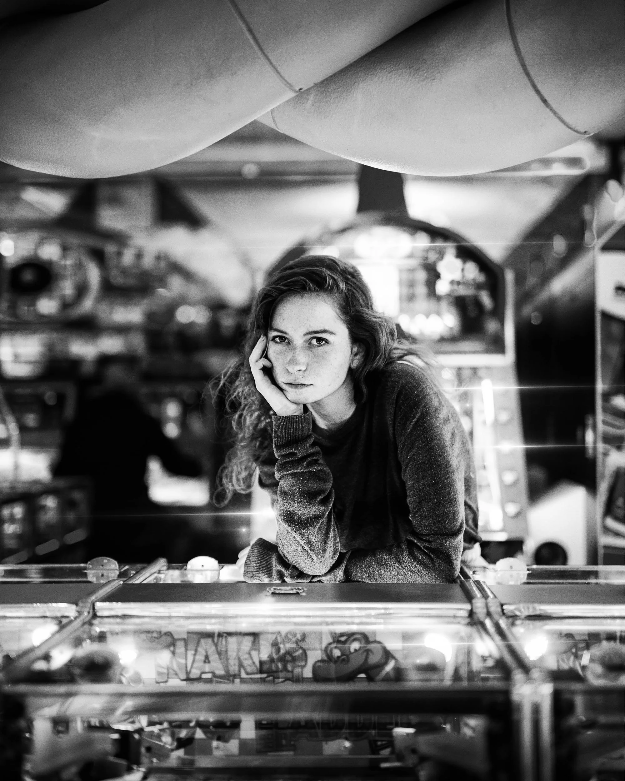 A young woman with curly hair and freckles leans on a pinball machine, resting her head on her hand and looking at the camera in a game arcade, black and white photo.
