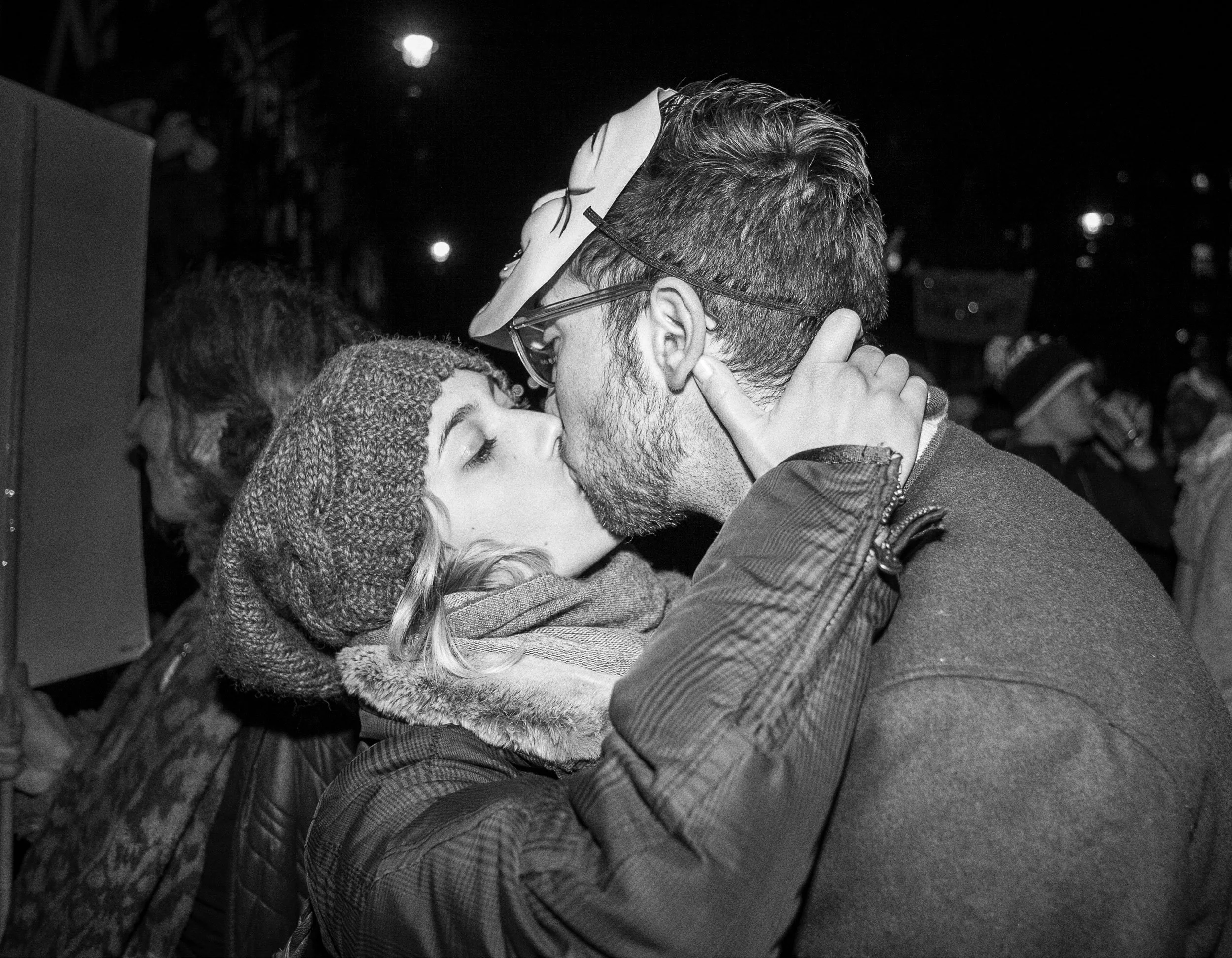 A couple kissing at night, with other people in the background.