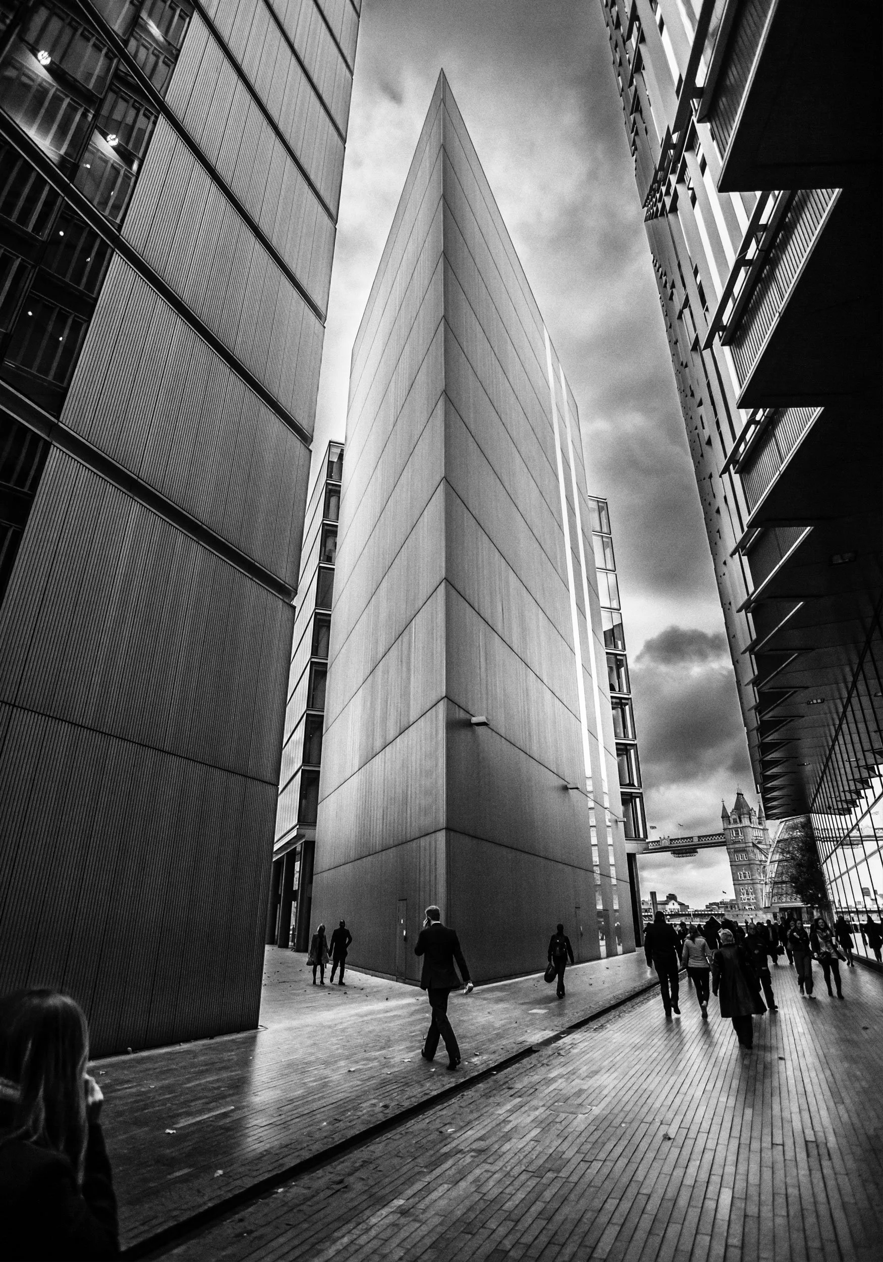 A black and white photo of tall, modern office buildings with glass and metal facades, with pedestrians walking on a wooden boardwalk and Tower Bridge visible in the background in London.