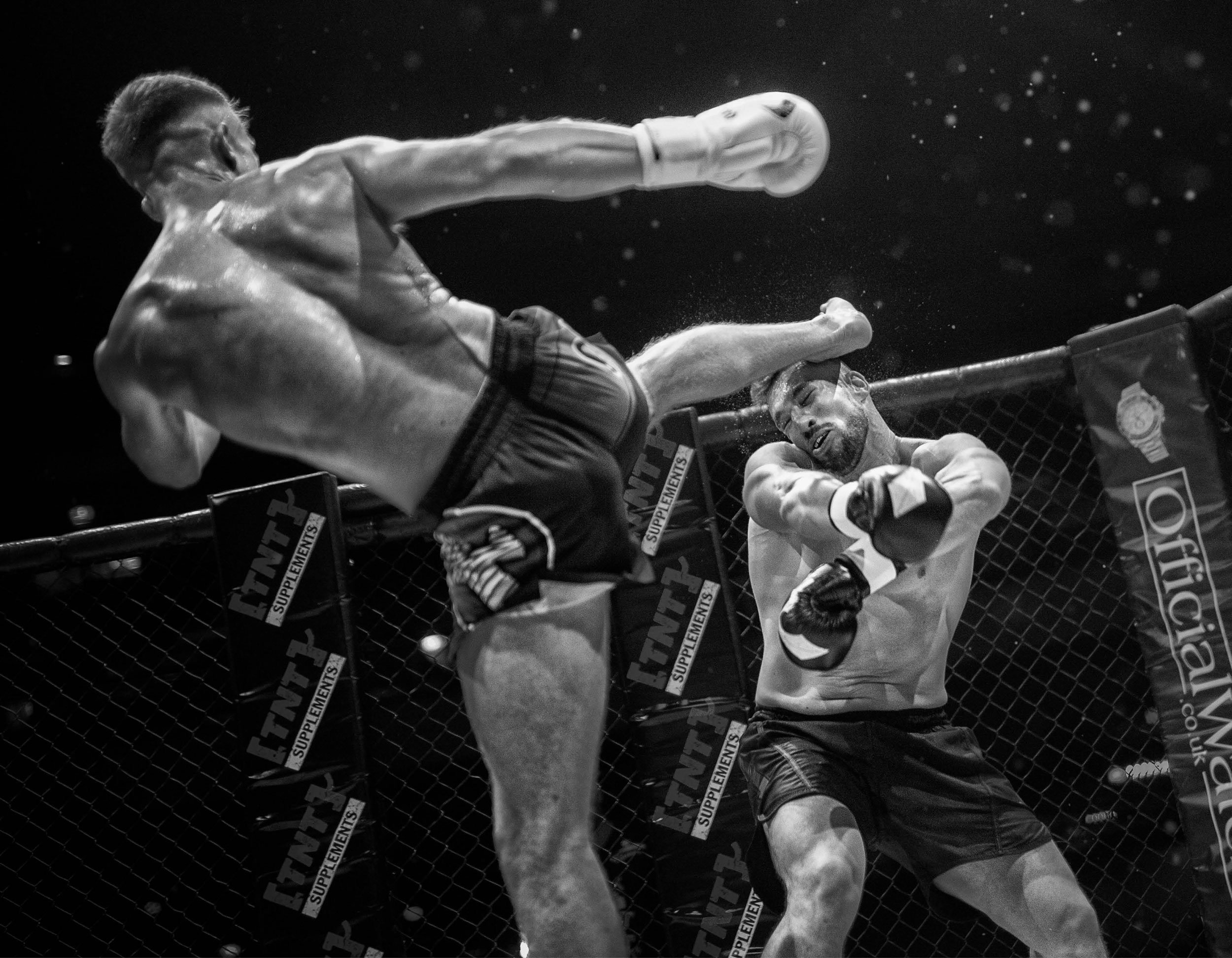 Black and white photo of two mixed martial arts fighters in a cage. The fighter on the left is executing a high kick towards the face of the fighter on the right, who is blocking the kick with his arms. Both fighters are shirtless, wearing gloves, an