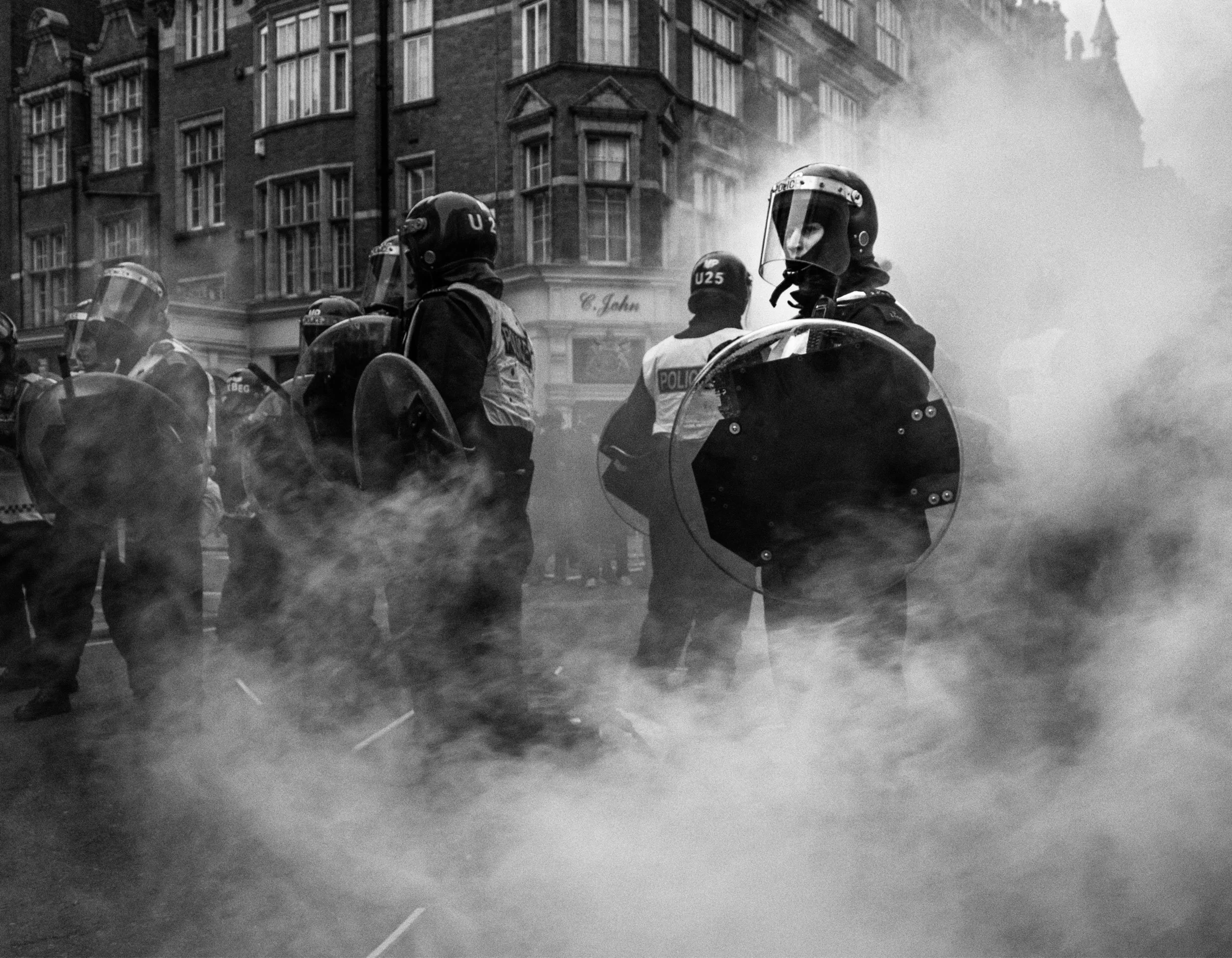 Police officers in riot gear holding shields and standing amidst smoke or tear gas, with urban buildings in the background.