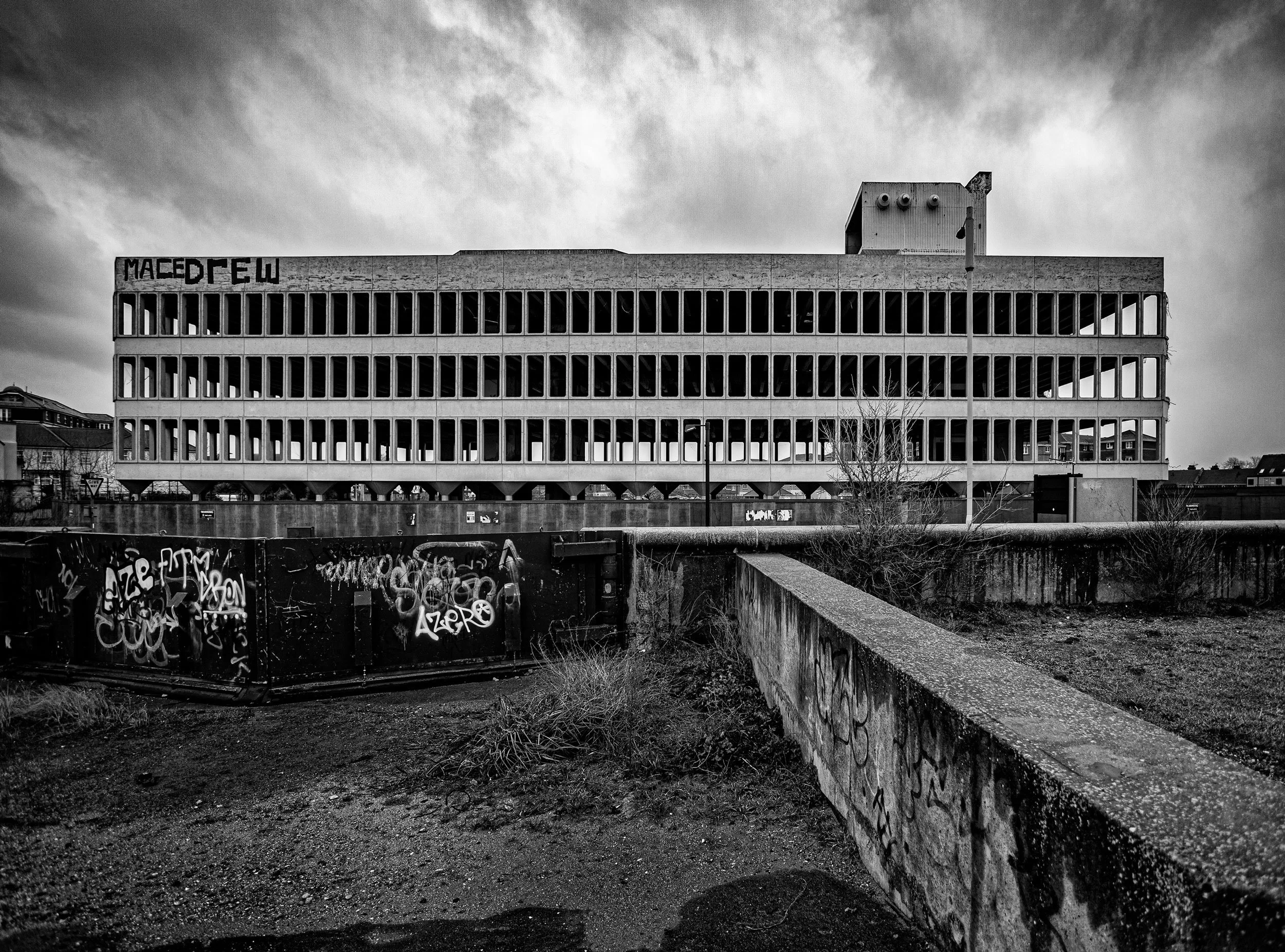 Black and white photo of an abandoned, graffiti-covered building with broken windows, seen from an empty foreground area with graffiti on a wall and a dirt ground. Old abandoned building in Southend, now demolished 