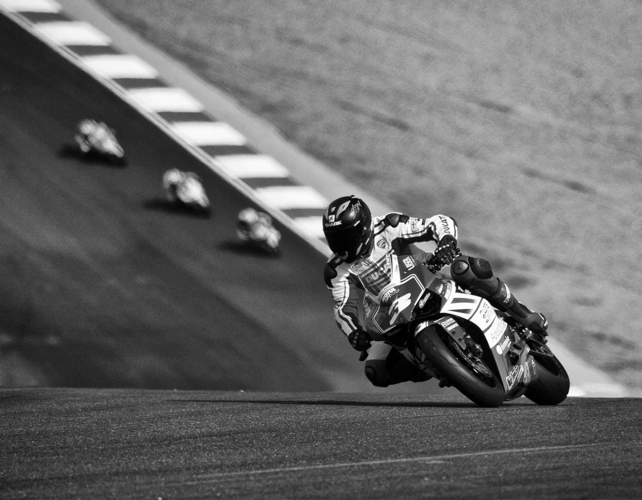Black and white photo of a motorcycle racer leaning into a turn on a racetrack, with several other racers visible in the background.
