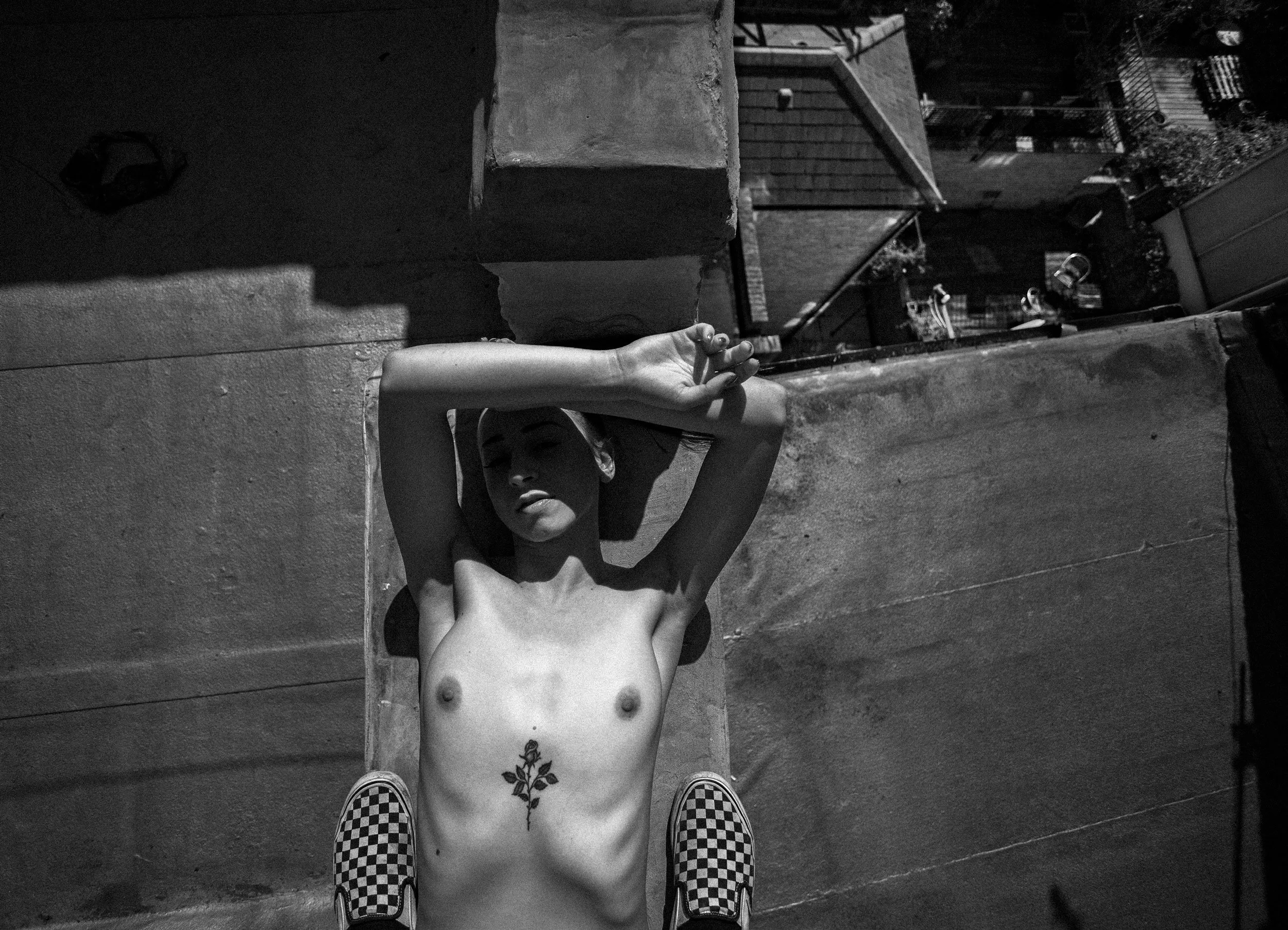 A topless woman with a floral tattoo on her abdomen lying on her back on a rooftop, with arms raised and hands clasped behind her head, wearing checkered slip-on shoes, with city rooftops in the background.