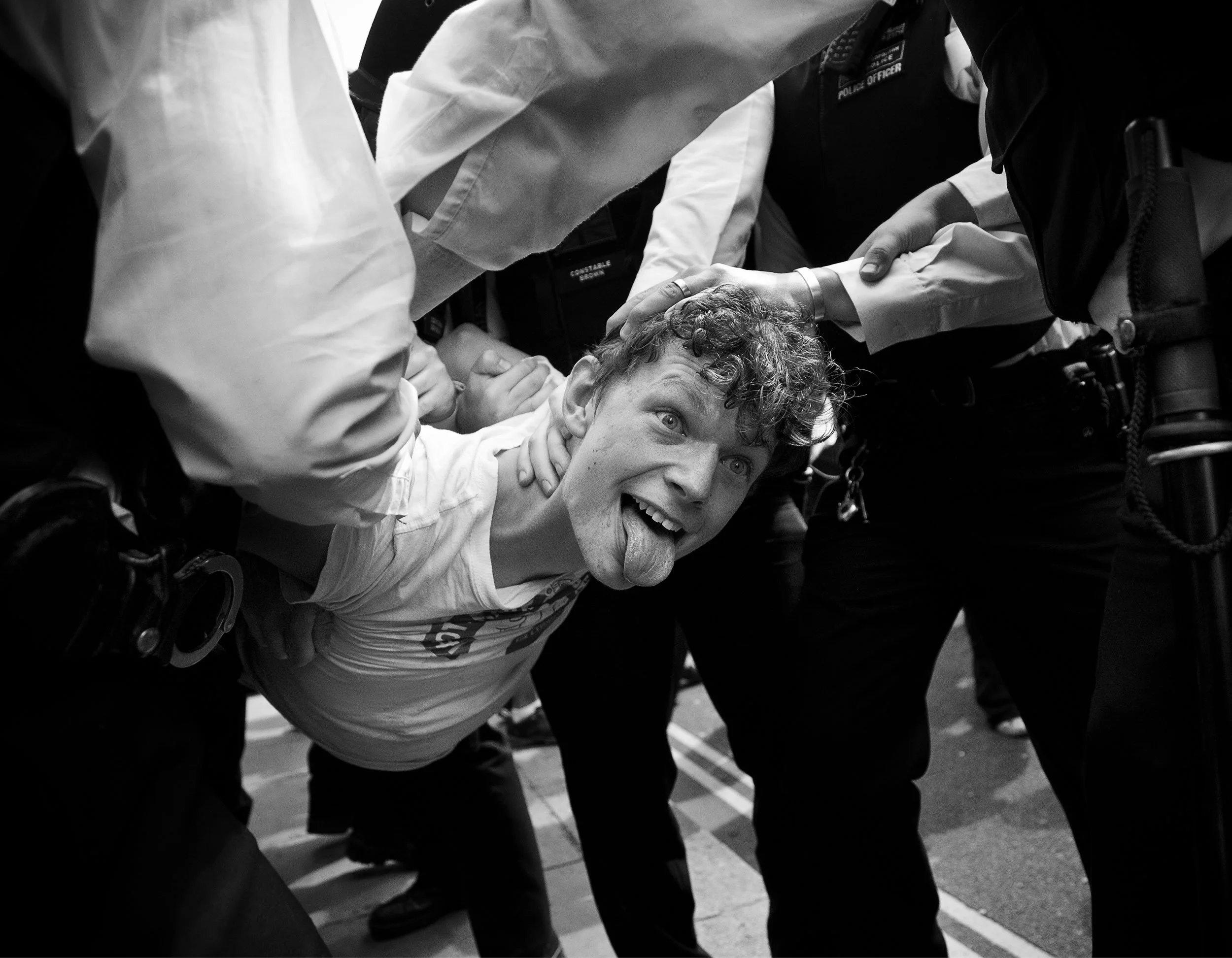 A young man with curly hair sticking out his tongue, being restrained by police officers during a protest.
