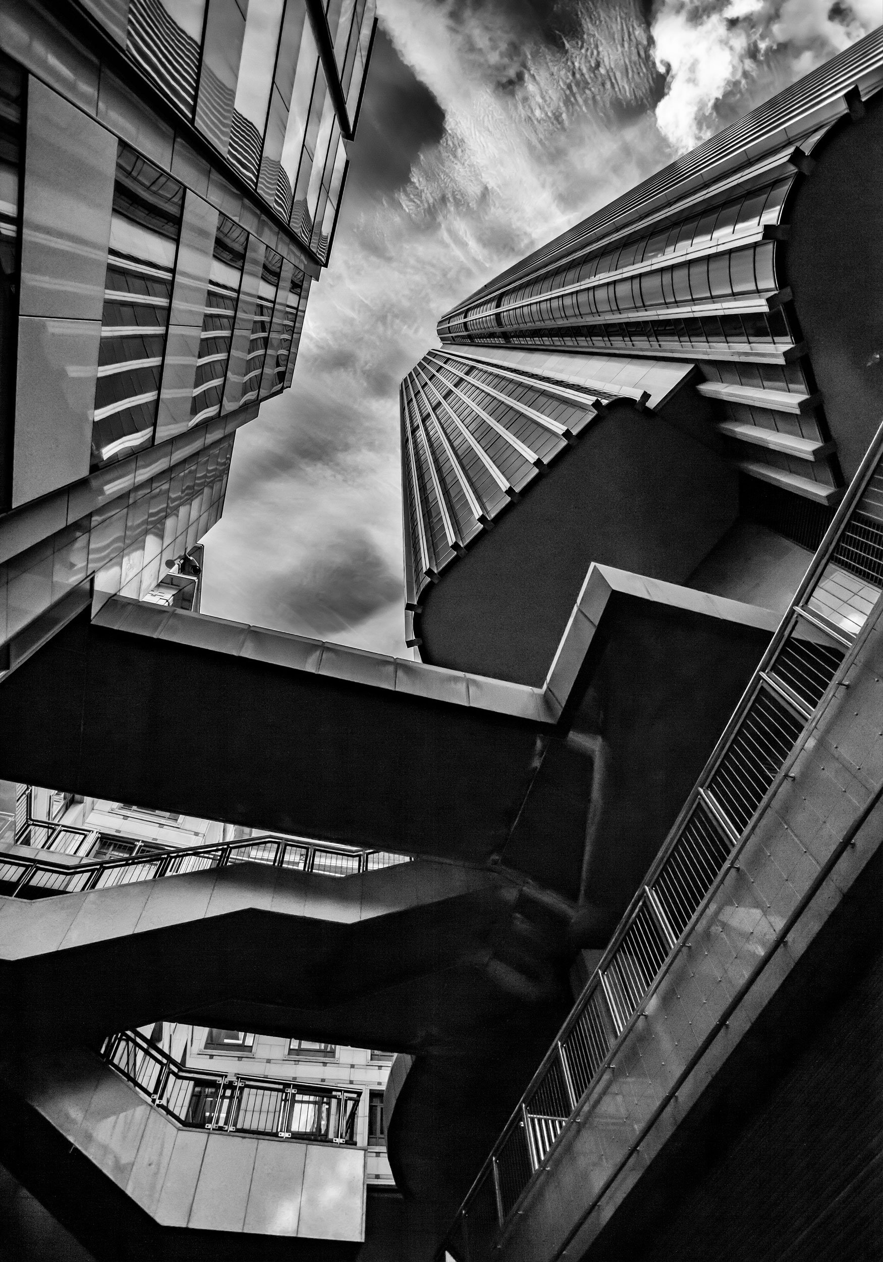 Looking up at modern high-rise buildings with glass facades and a network of outdoor walkways against a cloudy sky in black and white.