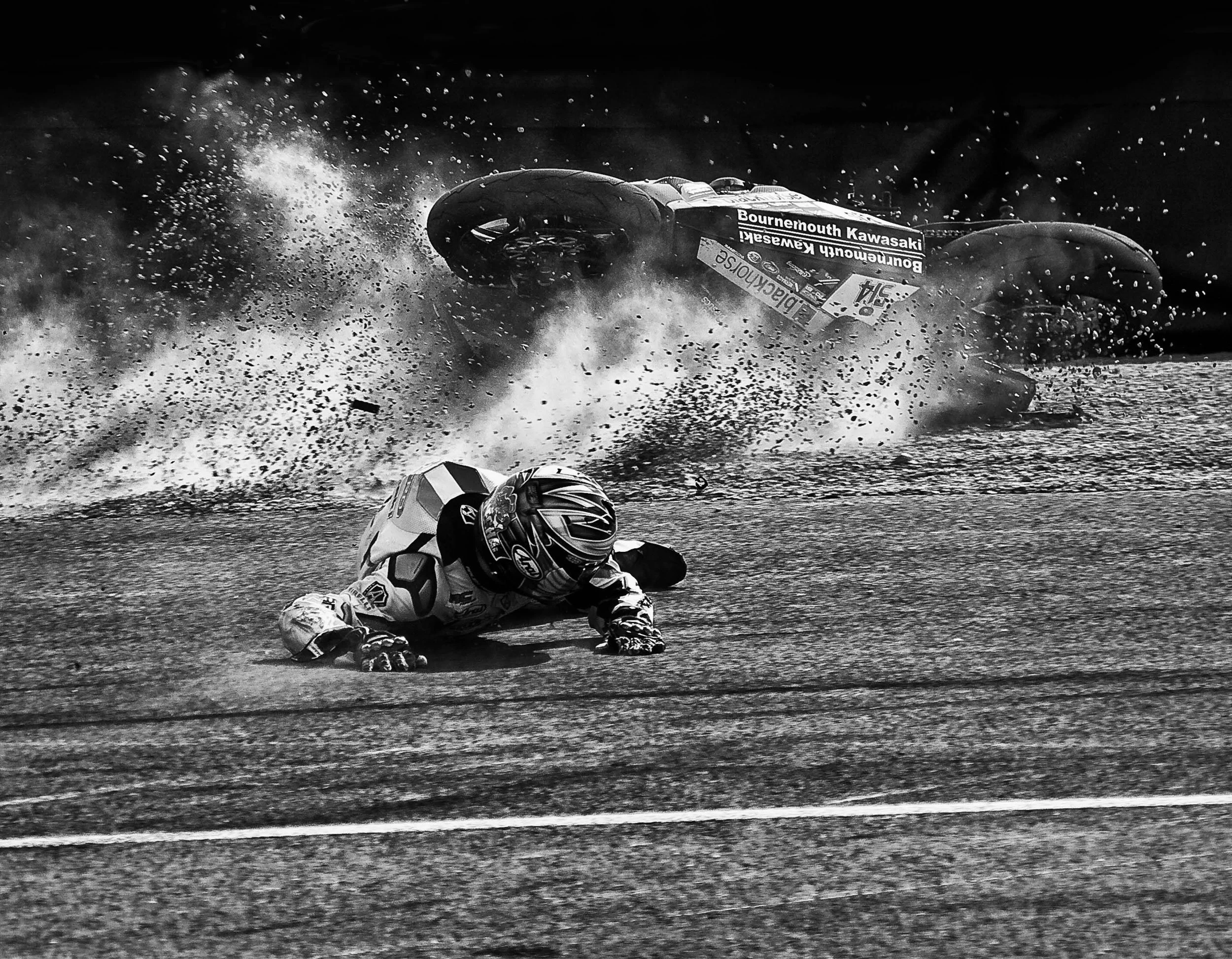 A motorcycle racer crashes, falling off the motorcycle into a water pool, with the motorcycle tumbling and water splash flying around in black and white.