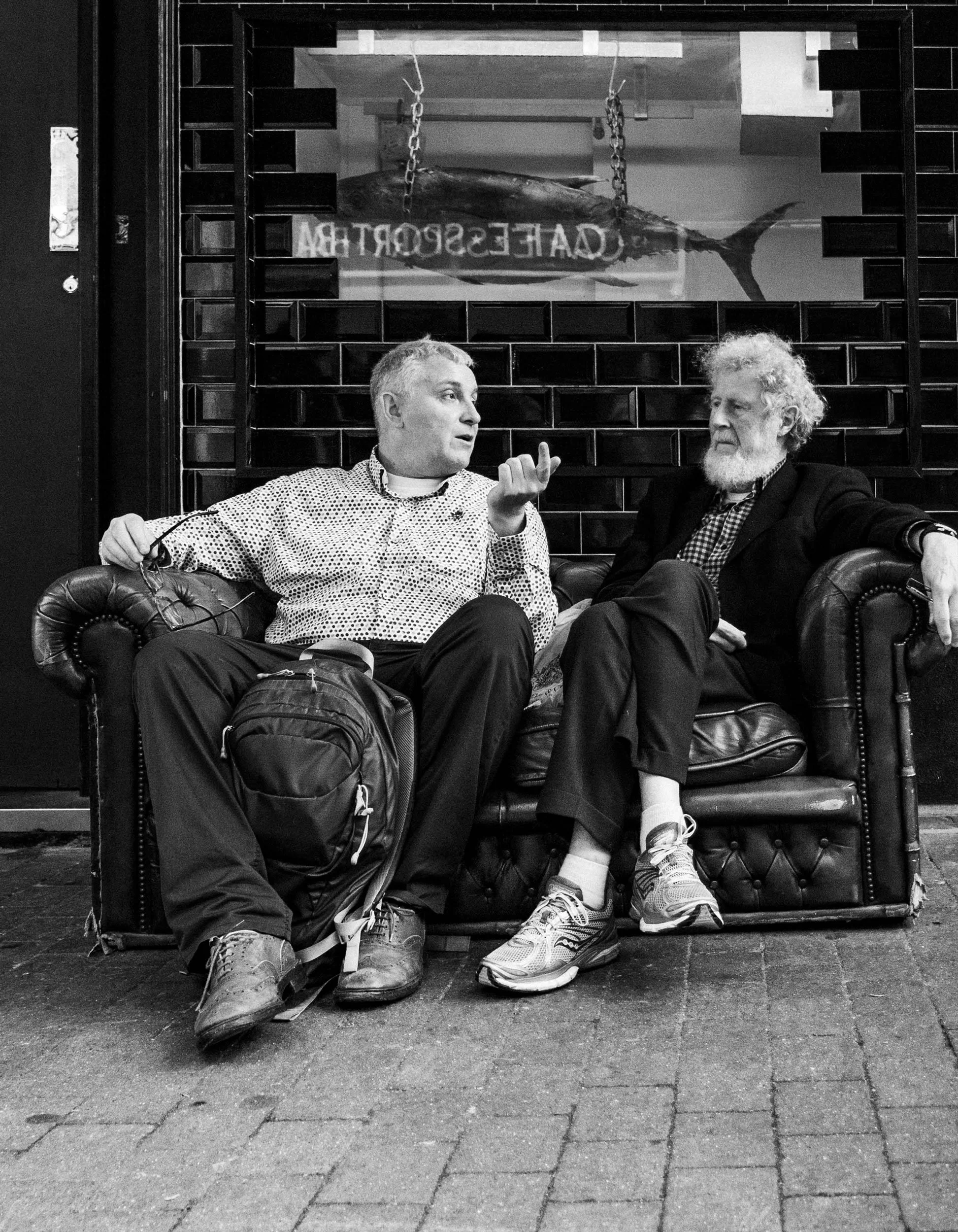 Two men sitting on a leather couch outside a restaurant, engaging in conversation. Behind them, there is a glass window with a fish sign and text that is mirror-reflected.