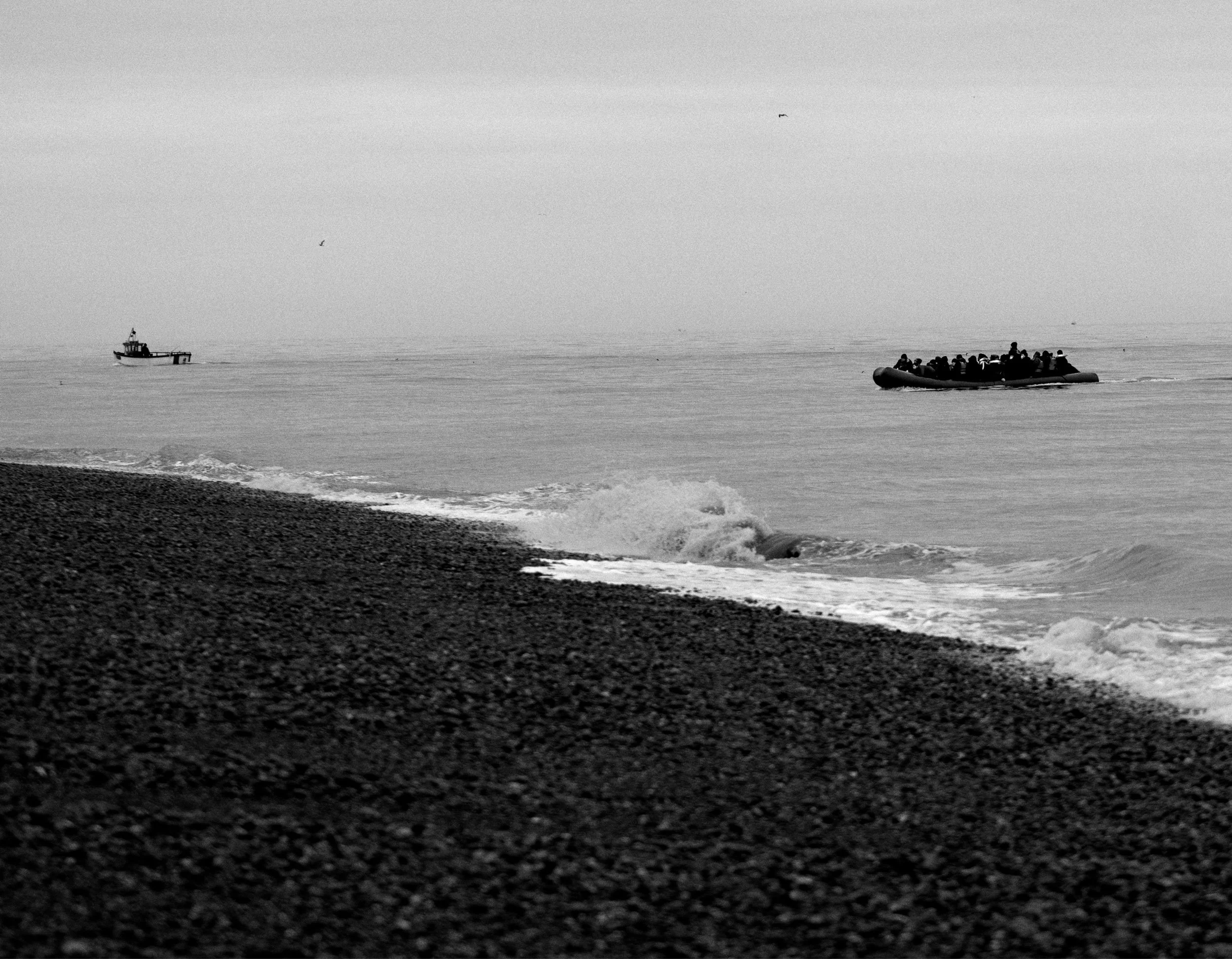 A black and white photo of a pebbled beach with waves approaching the shore. In the distance, a small boat is floating on the water to the left, and a larger rubber boat filled with people is moving across the water to the right.