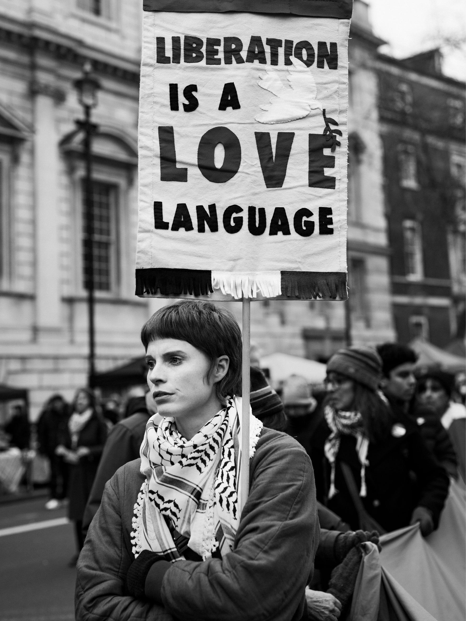 A woman with short dark hair and a striped scarf holds a protest sign that reads "Libertation is a Love Language." She is standing outdoors among a crowd of people, with historic buildings in the background.