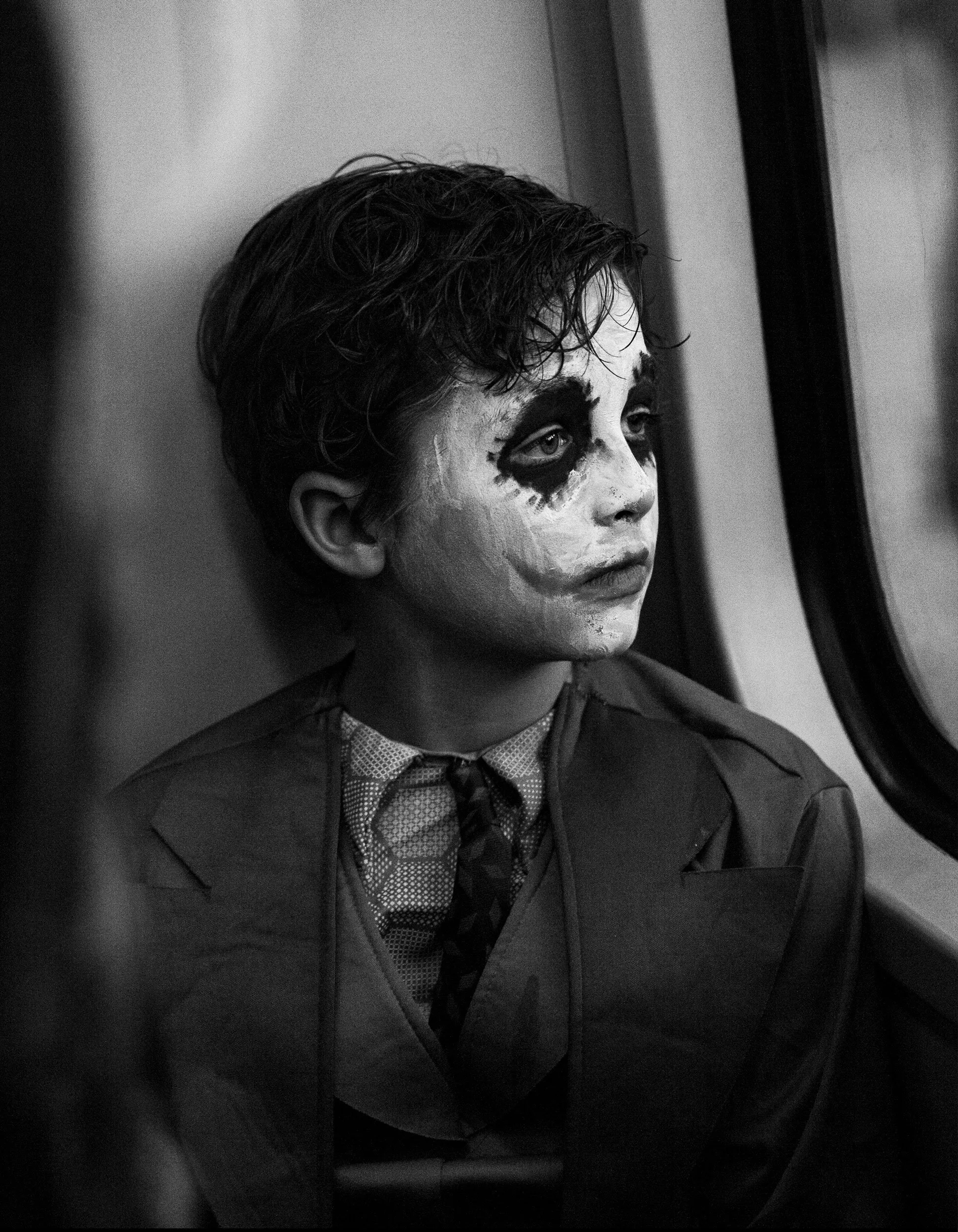 A person on a train or bus with face painted as a clown, looking out the window.