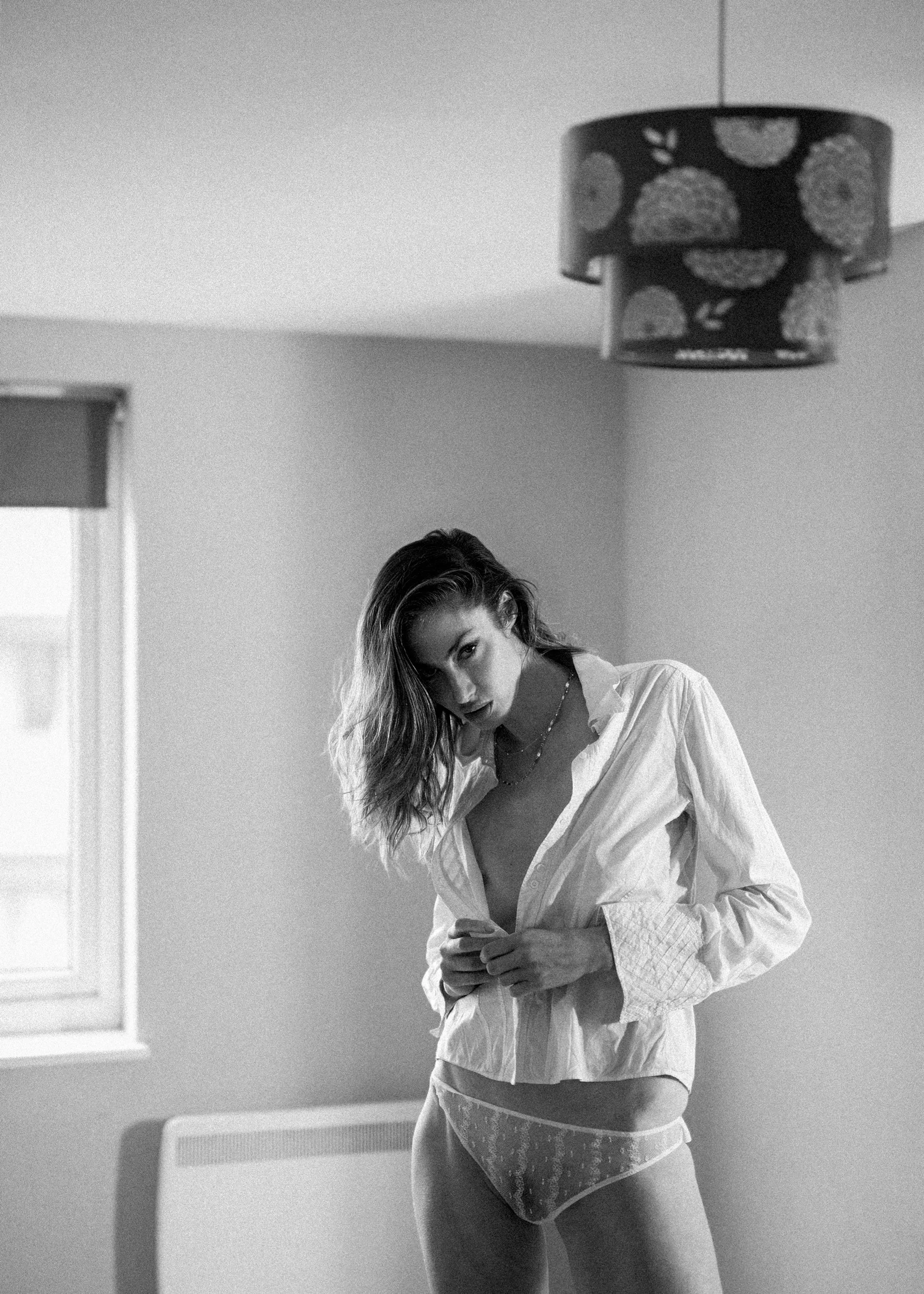 Black and white photo of a woman standing in a room, wearing a partially open shirt and lace underwear, looking at the camera with tousled hair.