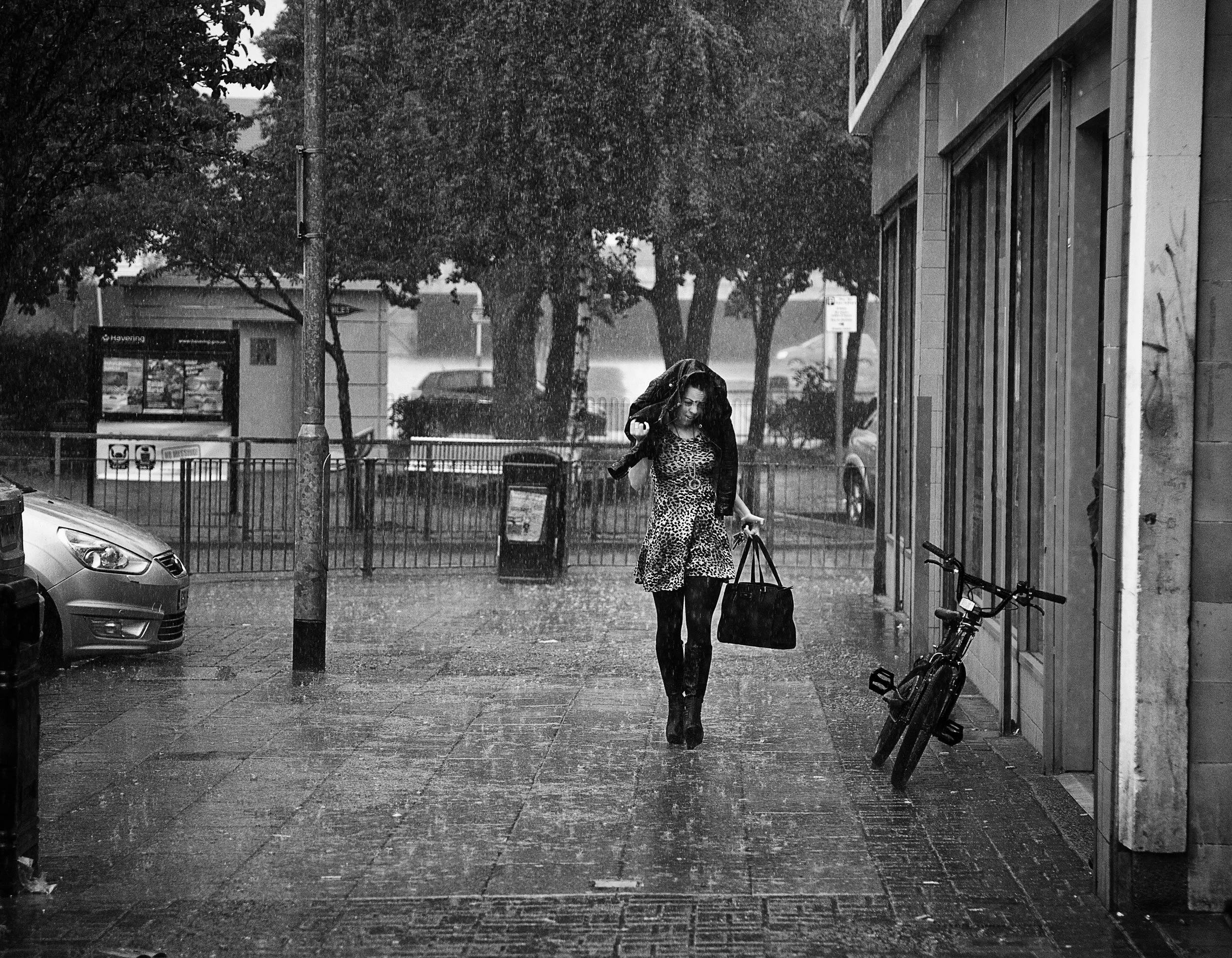 A woman wearing a leopard print dress and black tights walks in the rain while holding an umbrella. She carries a handbag and is near a bicycle on a city sidewalk.