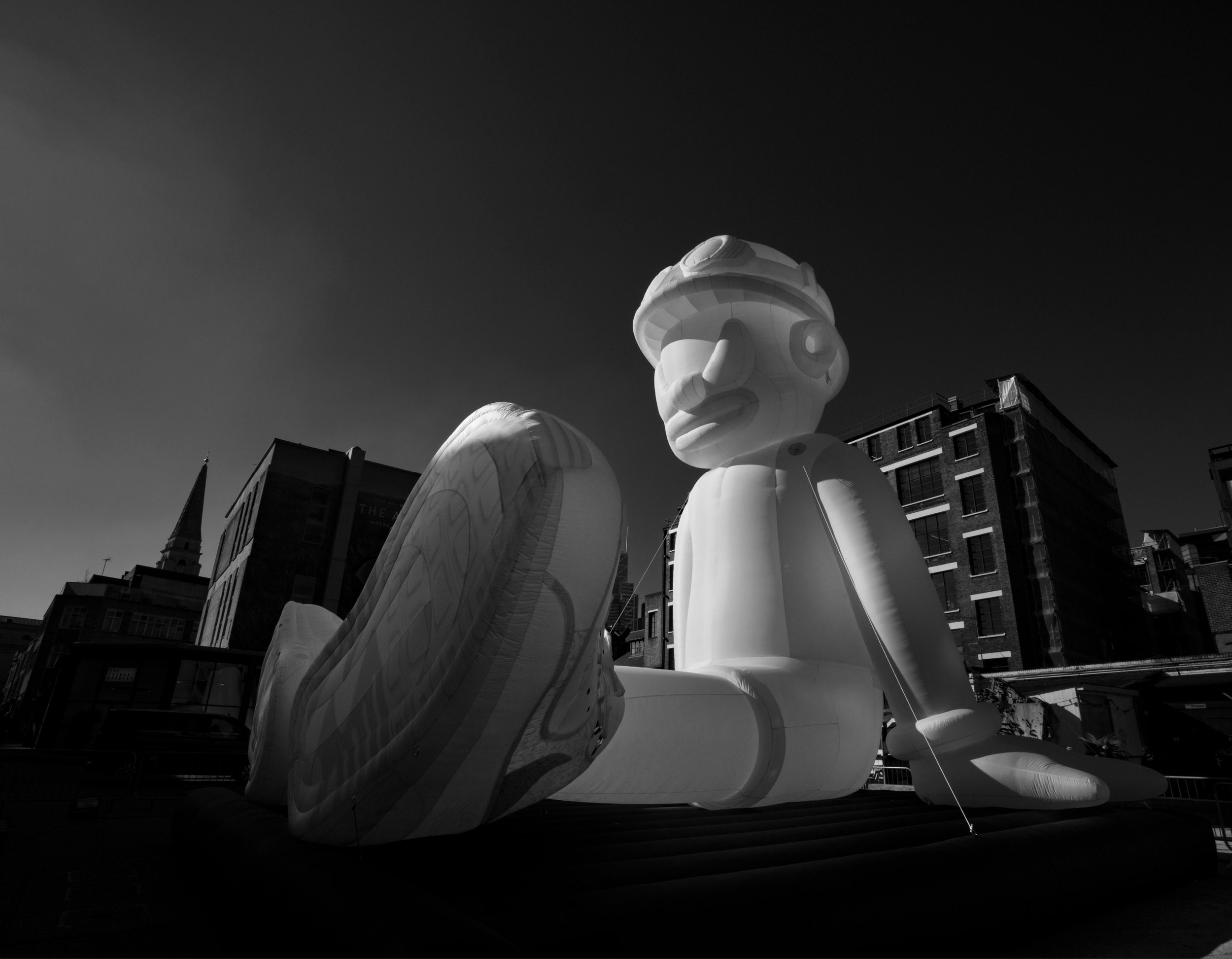 Large inflatable statue of a cartoon character with a large nose, wearing goggles and a cap, sitting on the ground in an urban area with buildings around.