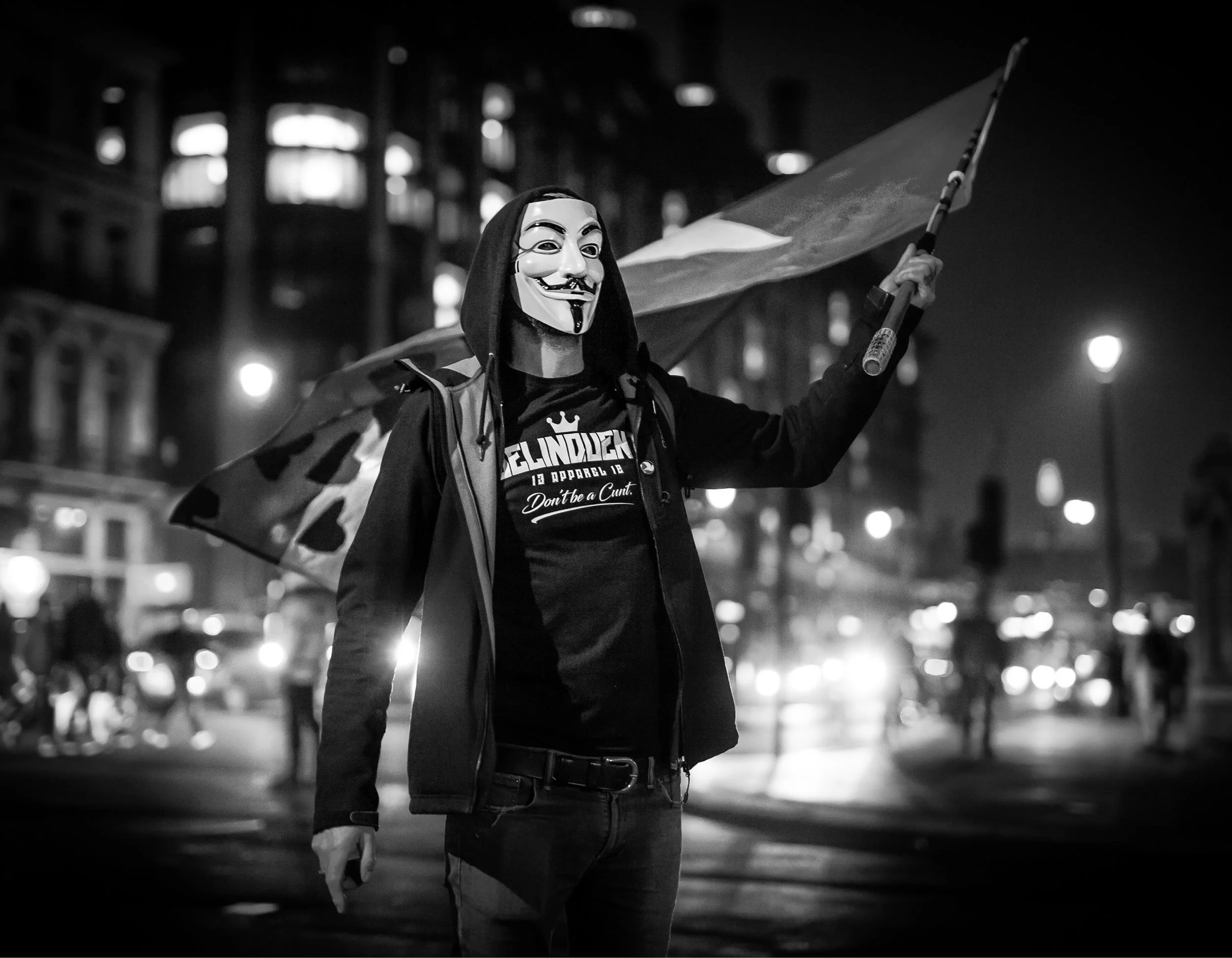 A person wearing a Guy Fawkes mask, hoodie, and t-shirt, standing in a city at night, holding a flag. The background shows blurred city lights and buildings.
