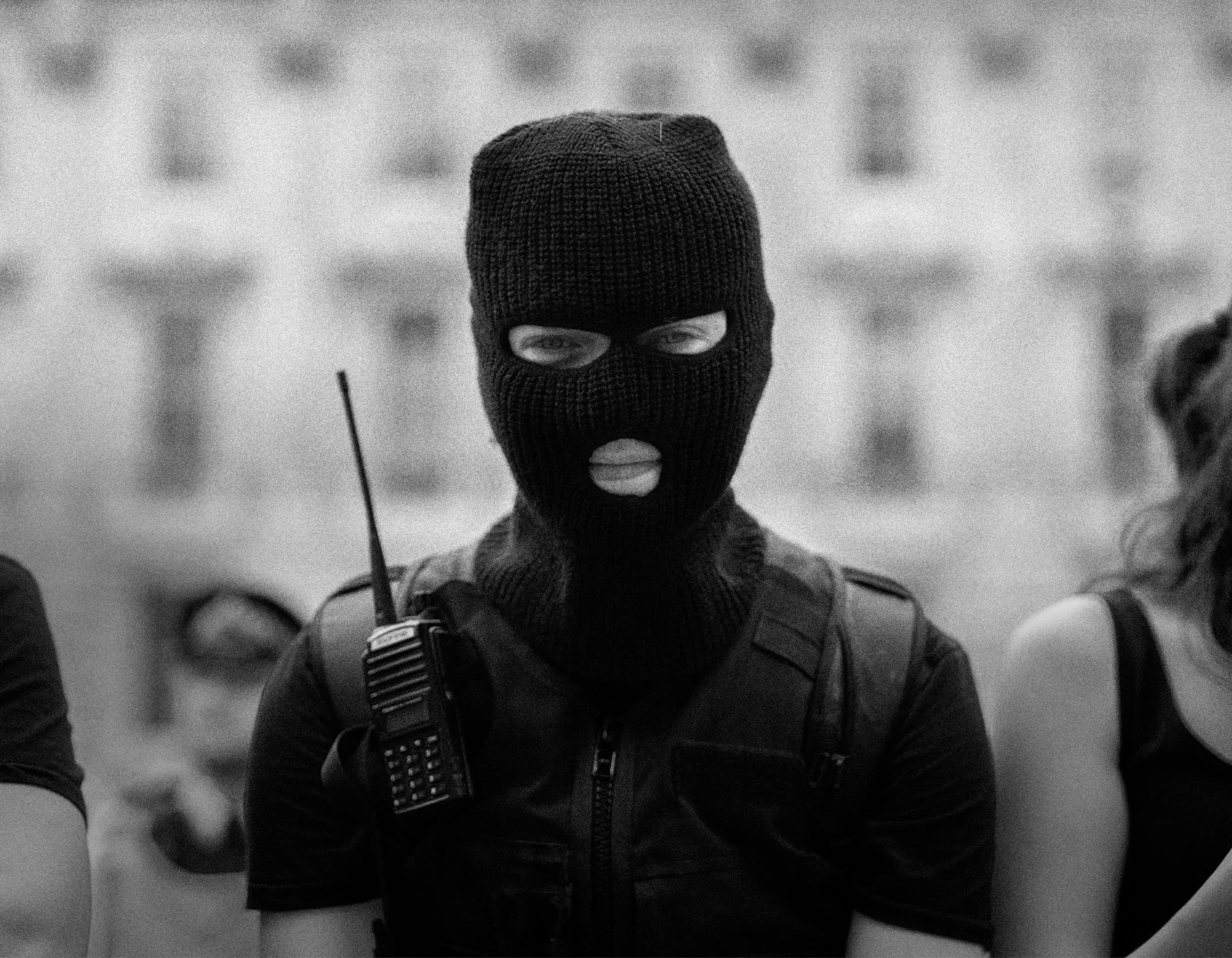 Person wearing a knit ski mask and tactical vest, with a walkie-talkie attached, posing outdoors in black and white.
