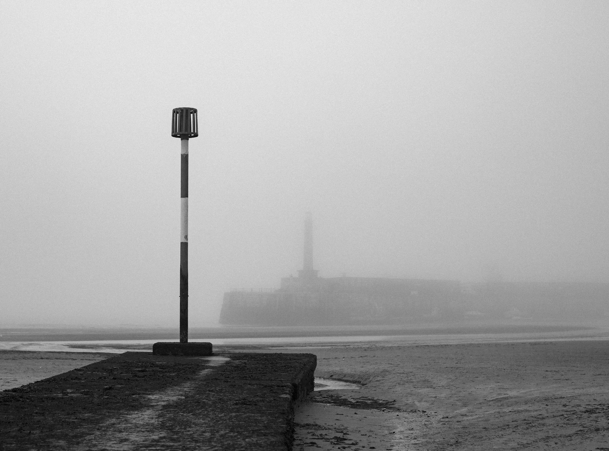 A foggy beach with a lone lamppost on a dock in the foreground and the silhouette of an old industrial building with a tall chimney in the distance. Margate beach in the fog