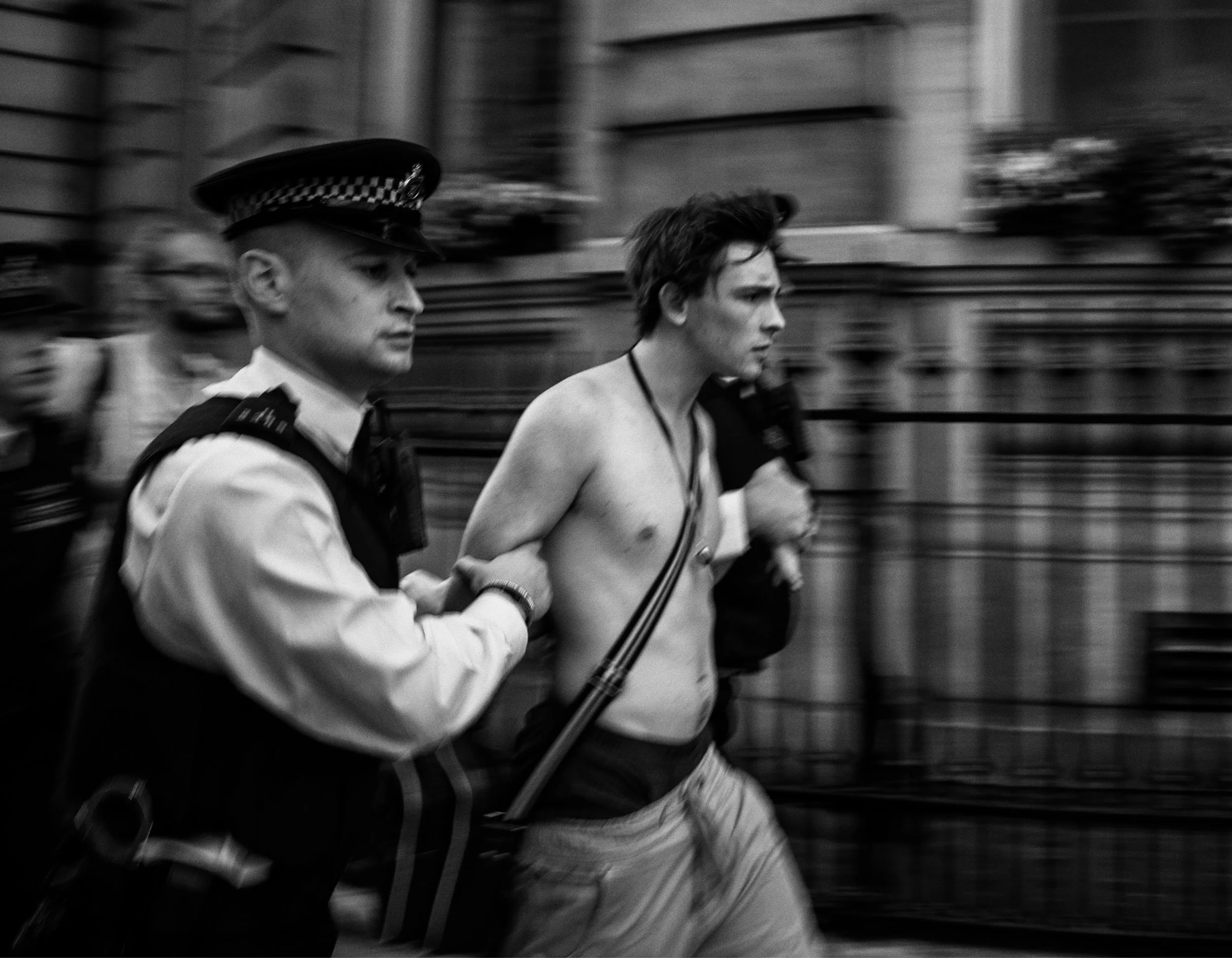 A black and white photo of a shirtless young man being escorted by a police officer.