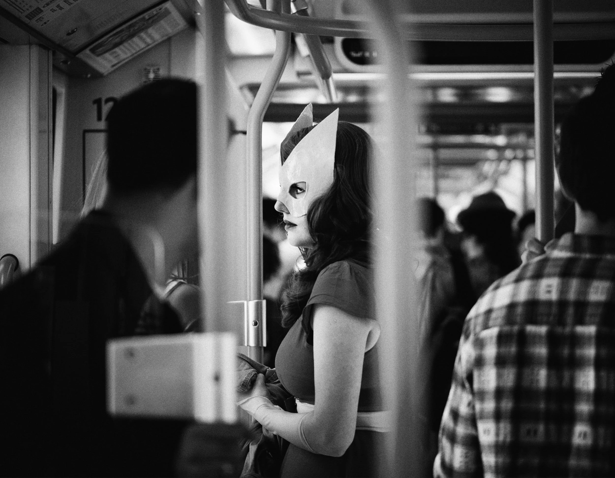 A woman wearing a cat-like mask, standing on a crowded train, surrounded by passengers, in a black and white photograph.