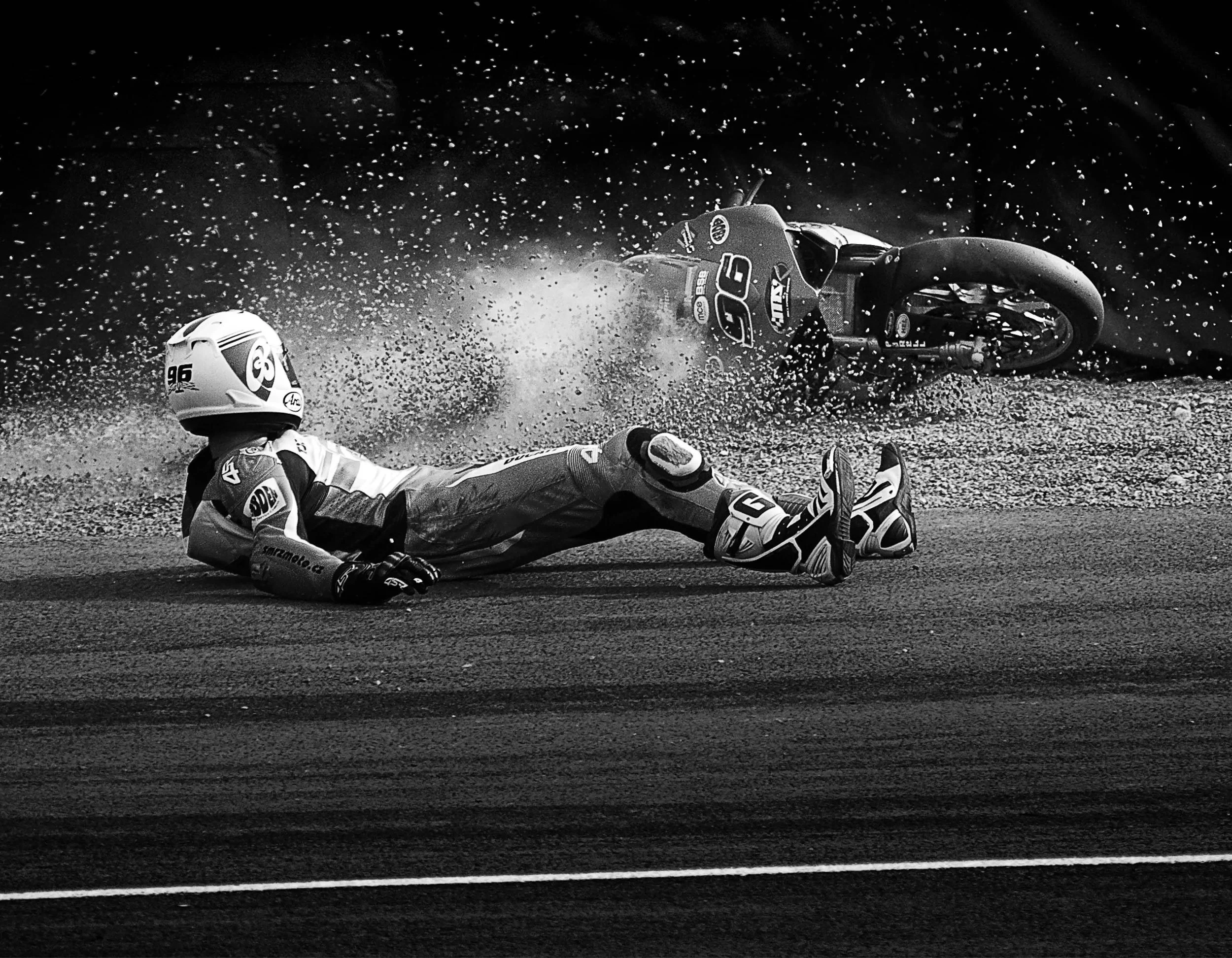Motorcycle rider falling off motorcycle during race, motorcycle skidding on track, rider lying on ground wearing helmet and protective gear, black-and-white photo.