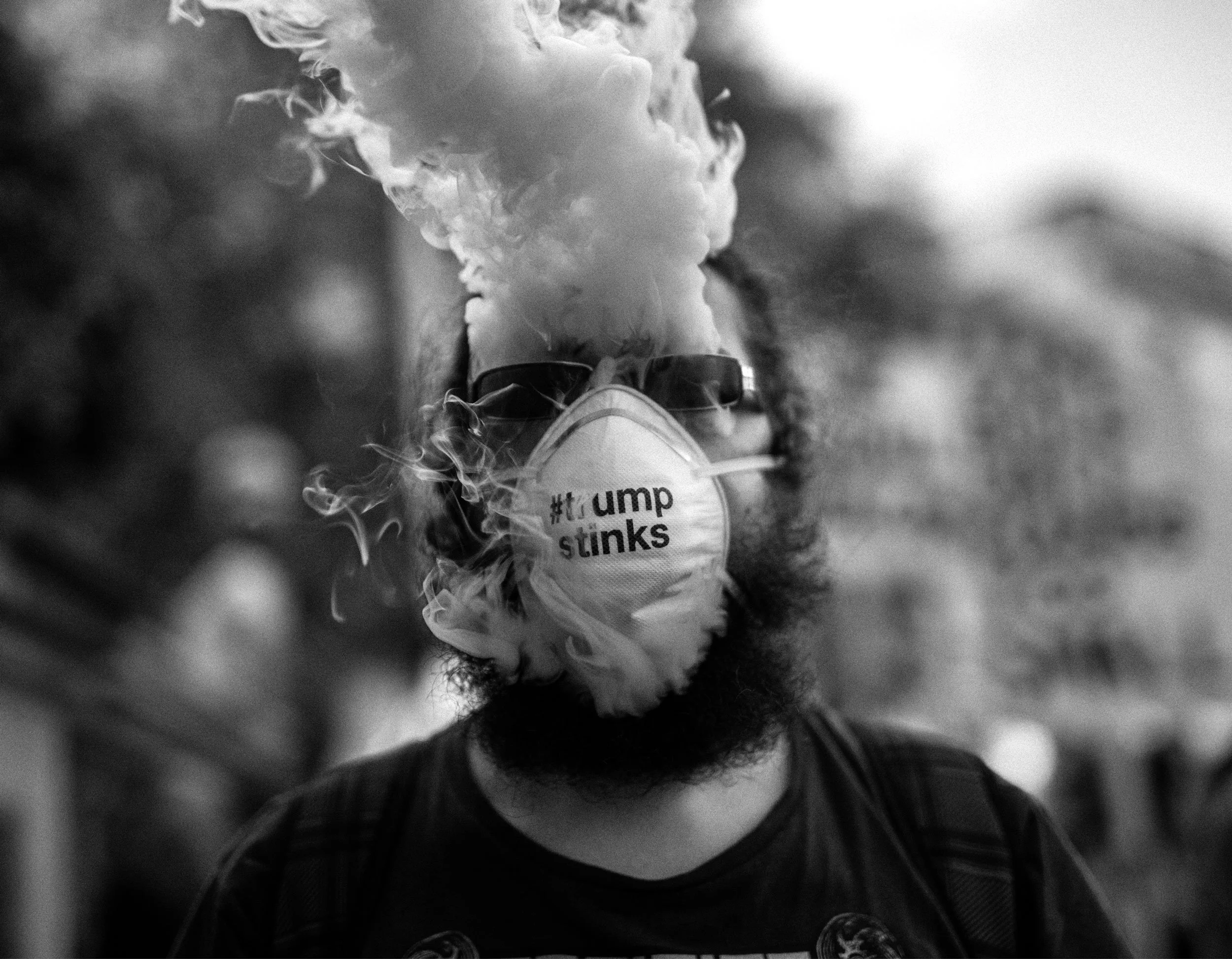 A person wearing sunglasses and a face mask with hashtag '#tRump stinks' on it, exhaling thick smoke or vapor outdoors.