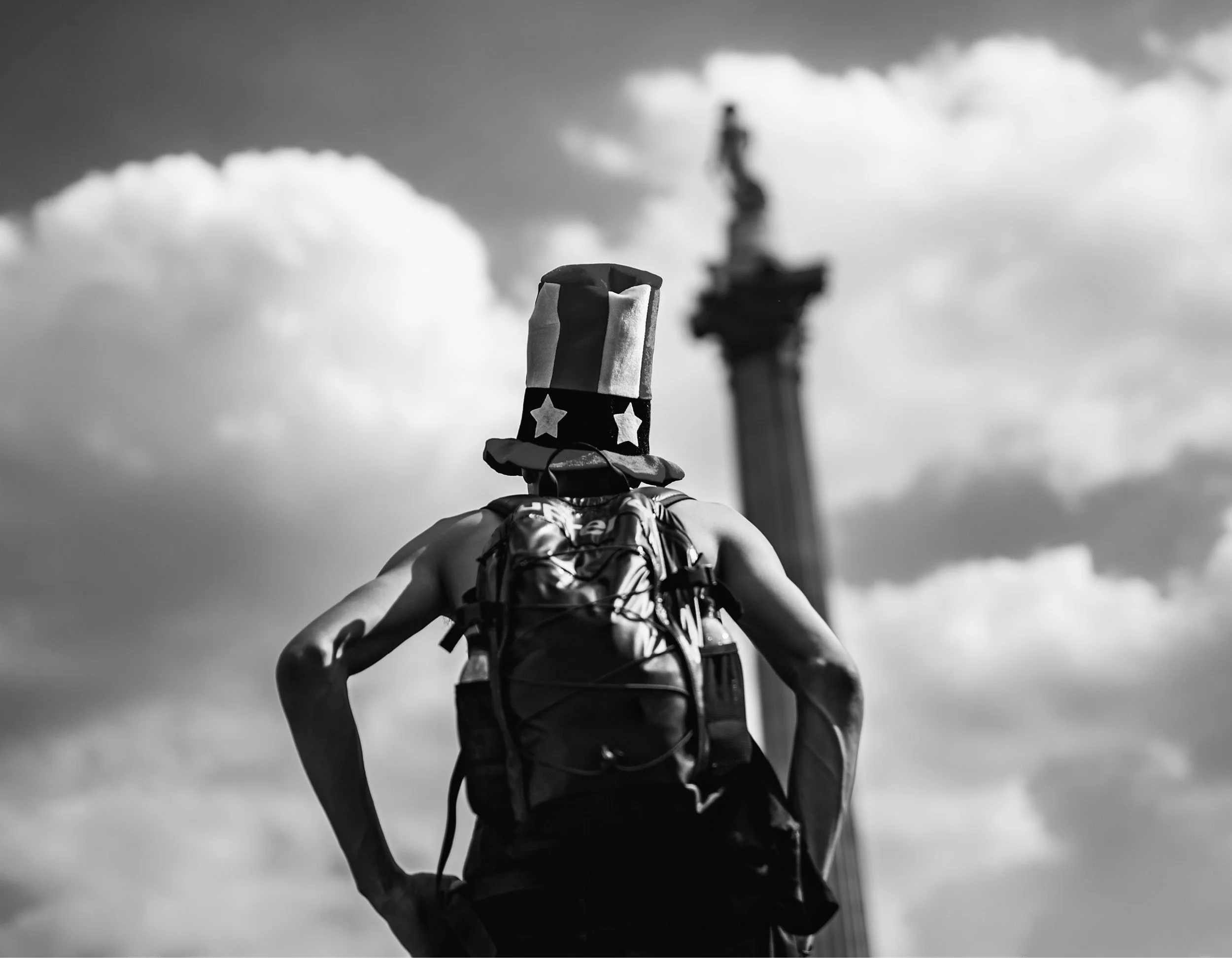 A person wearing a backpack and a tall hat with stars, facing away, near a monument with a statue on top, set against a cloudy sky.