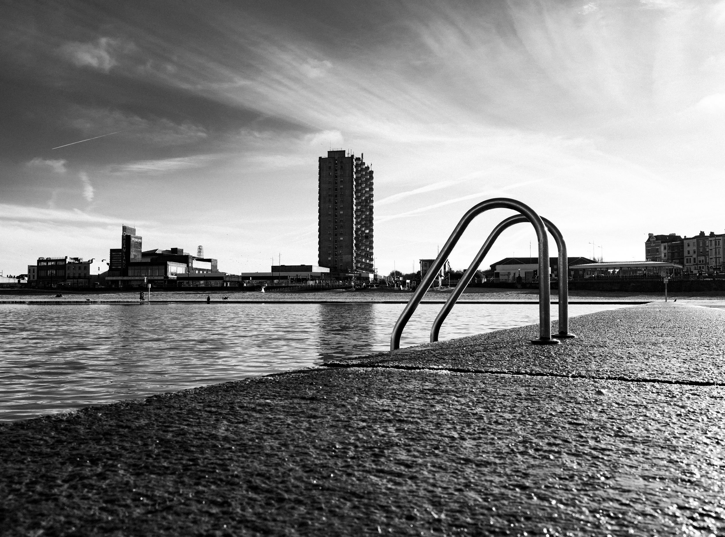 Black and white photo of a waterfront area with stairs leading into the water, buildings, and a tall skyscraper in the background under a partly cloudy sky.