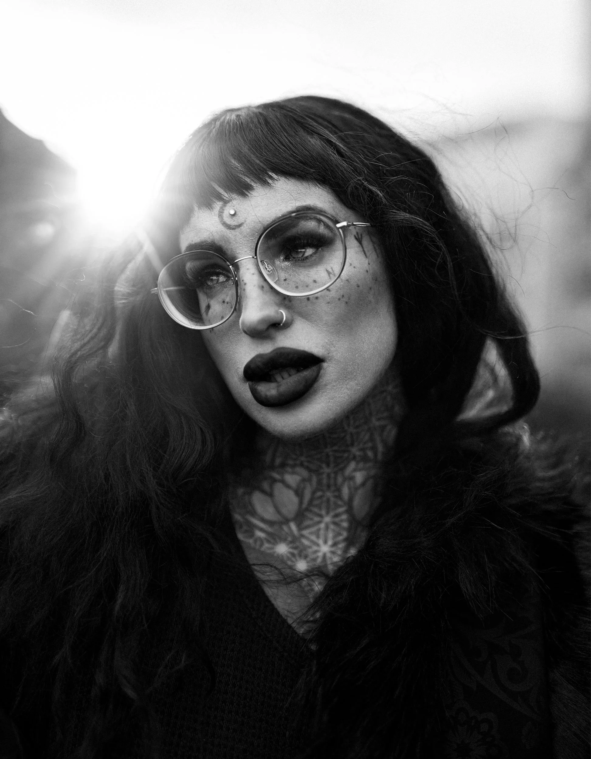 Black and white portrait of a woman with dark hair, tattoos, round glasses, and bold makeup, looking to the side with sunlight behind her.