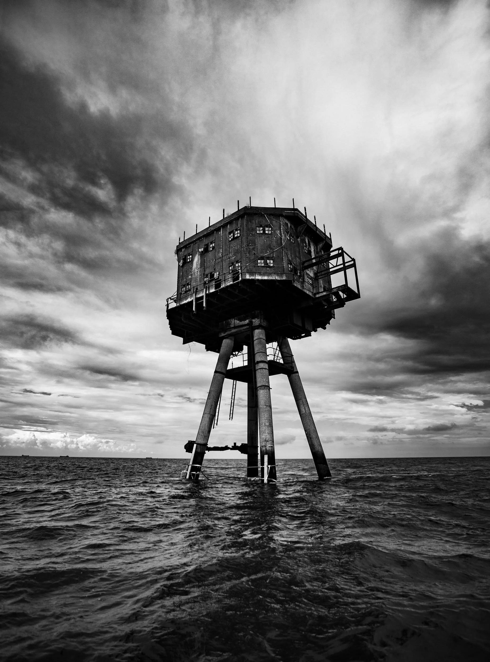 A black and white photo of an old, rusty offshore oil platform or lighthouse standing in the ocean with an overcast sky.