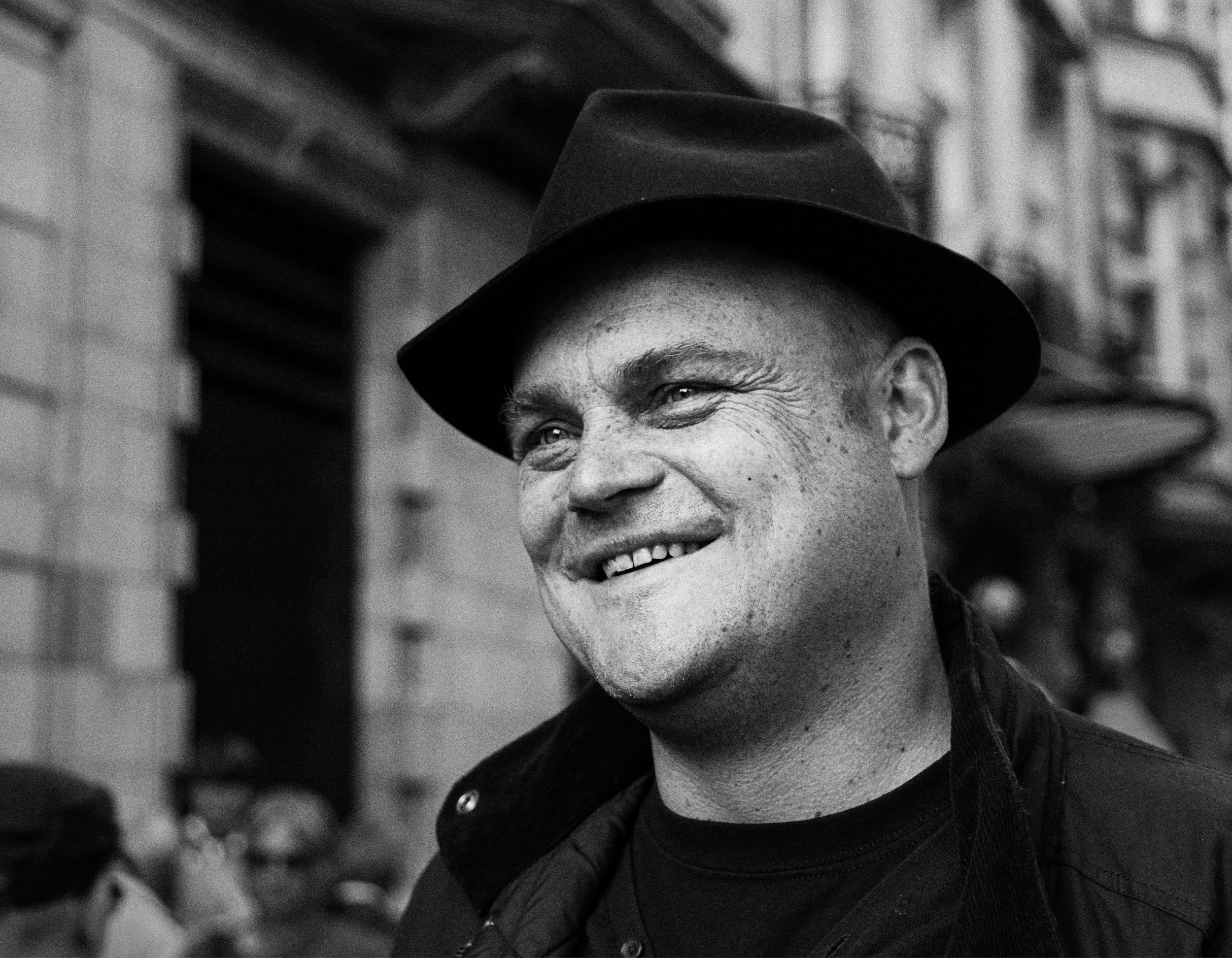 A black and white photo of a smiling man wearing a fedora hat and a dark jacket, with a brick building in the background.