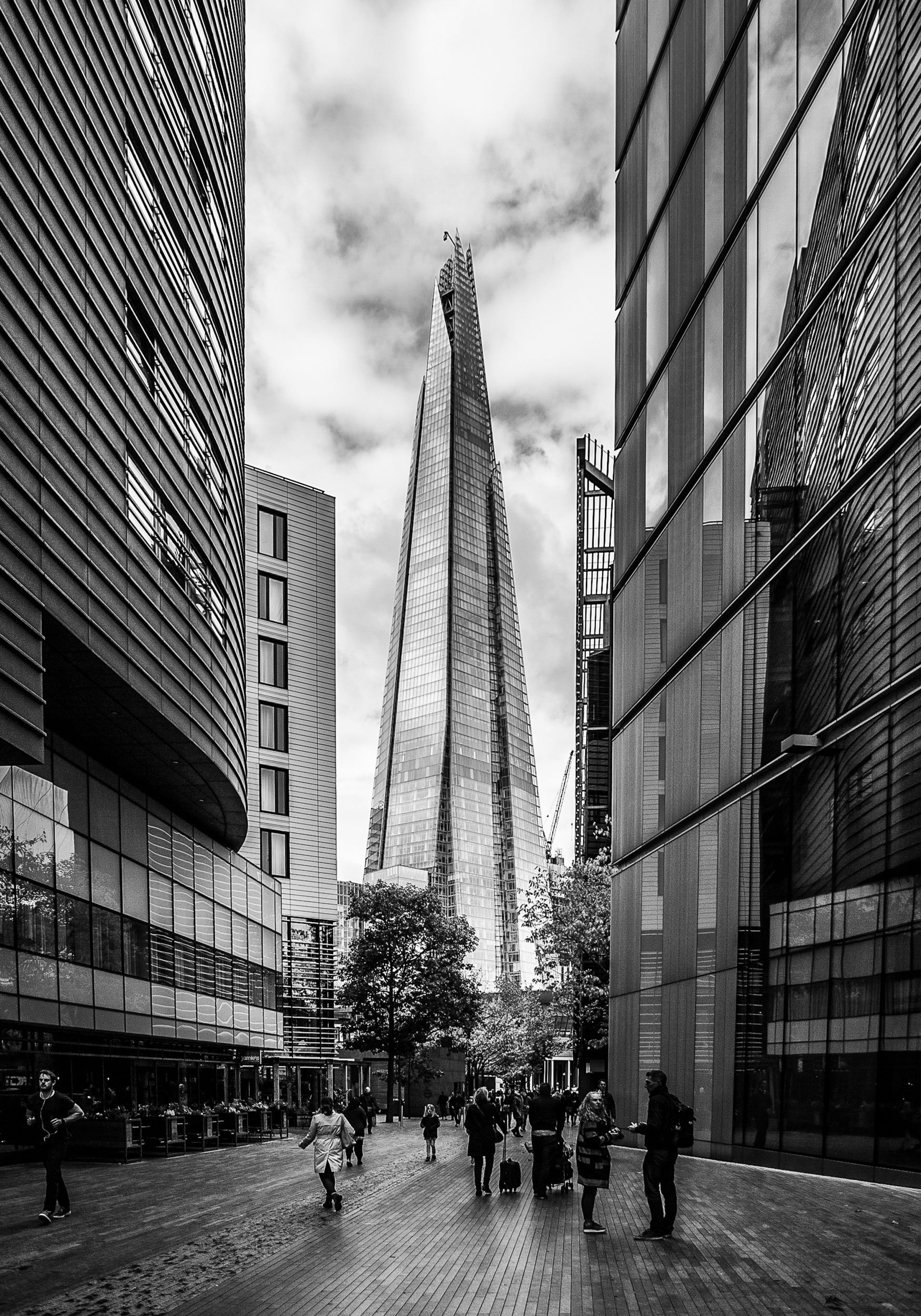 Black and white photo of a city street with tall modern buildings, people walking, and The Shard skyscraper in London in the background.