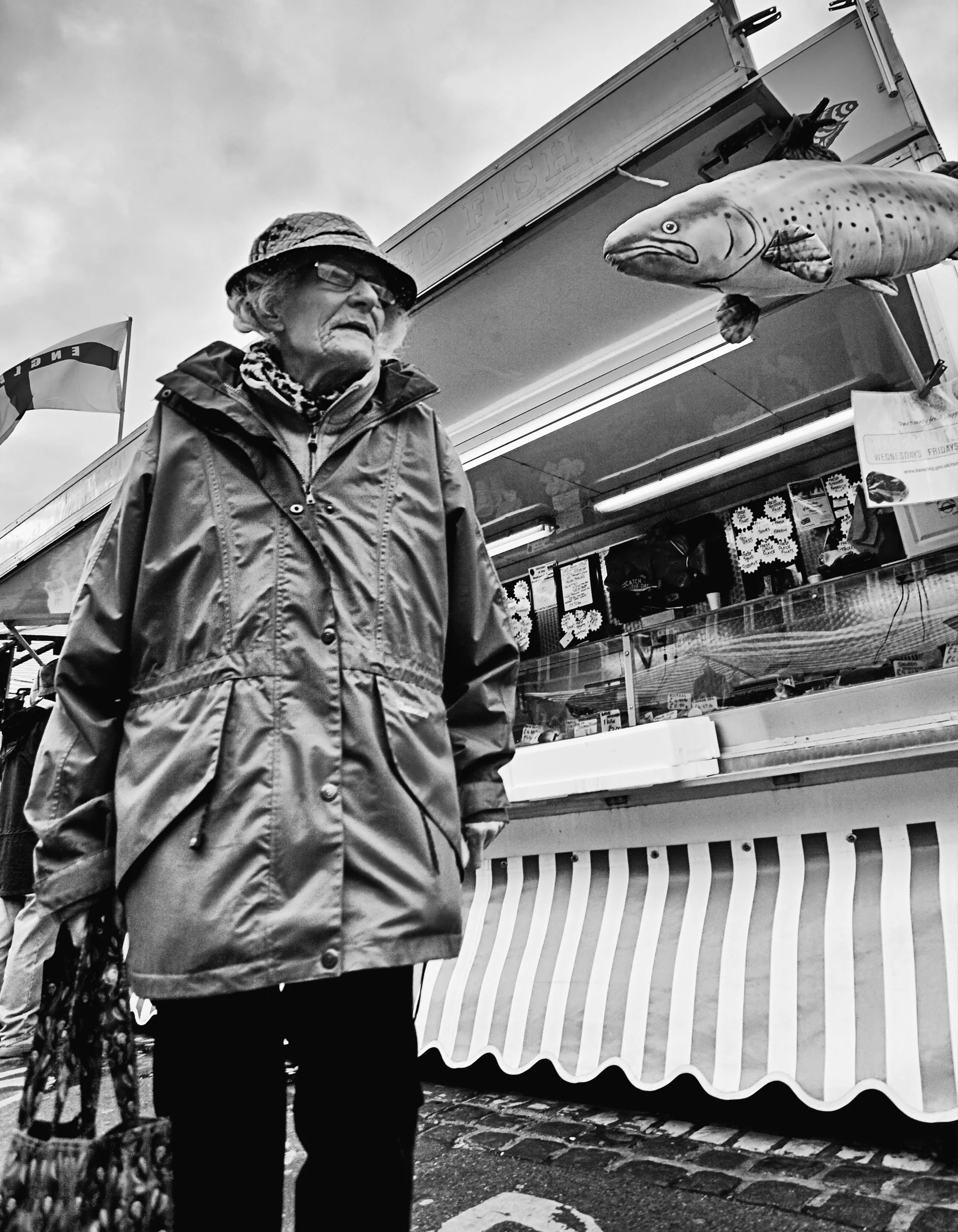 An elderly woman wearing a rain jacket, hat, and glasses standing in front of a fish stand at an outdoor market.