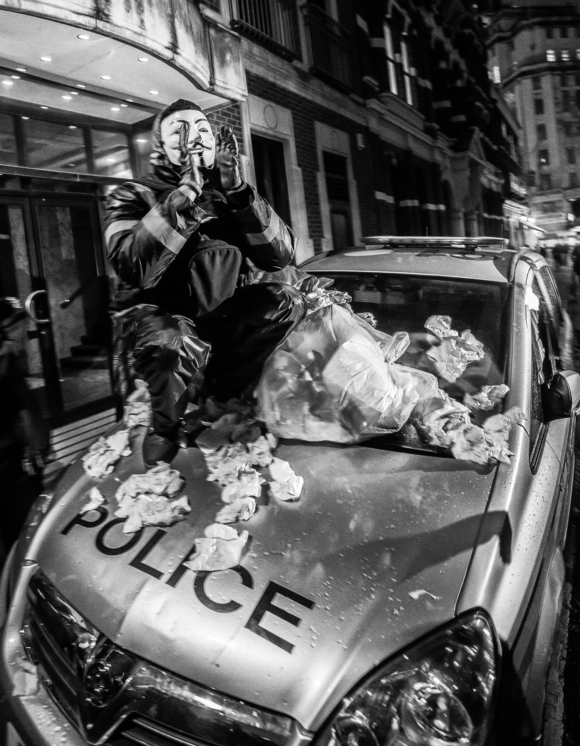 A person wearing a Guy Fawkes mask, sitting on a police car with crumpled paper around them and on the hood, in an urban street at night.