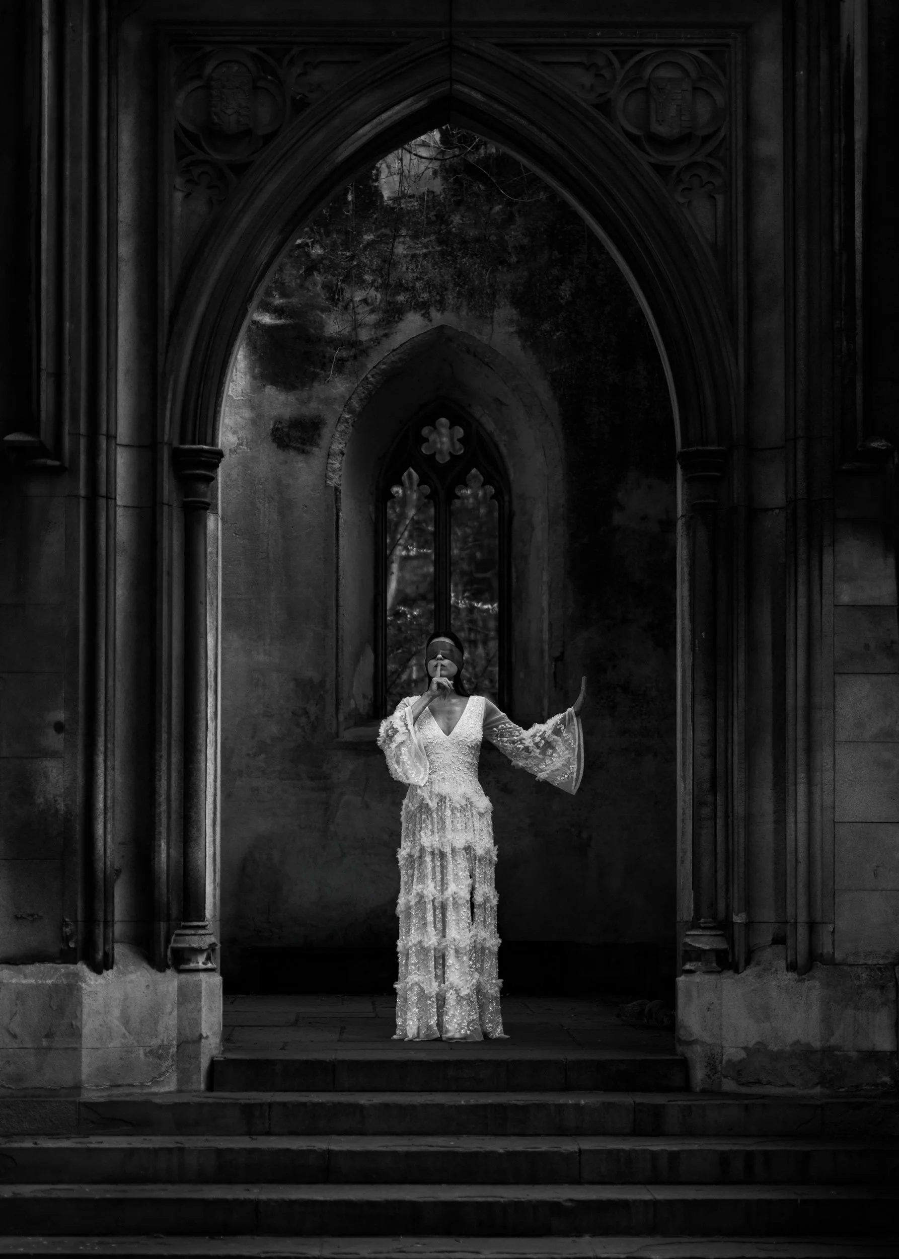 A woman in a long, ornate lace dress stands on steps in front of a Gothic-style arched window, with dark, weathered stone walls surrounding her, in black and white. Irida the Shapeshifter