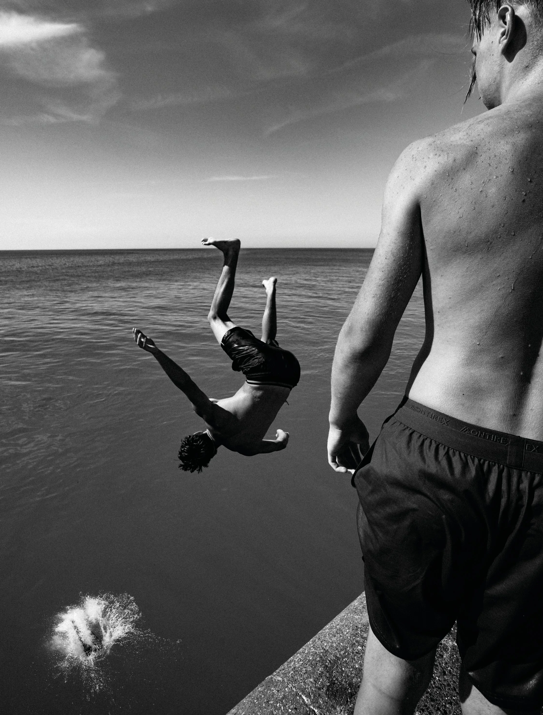 Margate harbour wall. A black and white photo of two young men at the edge of a body of water, one standing and observing, the other mid-fall after jumping, with water splashing as he enters the water from a dock or pier.