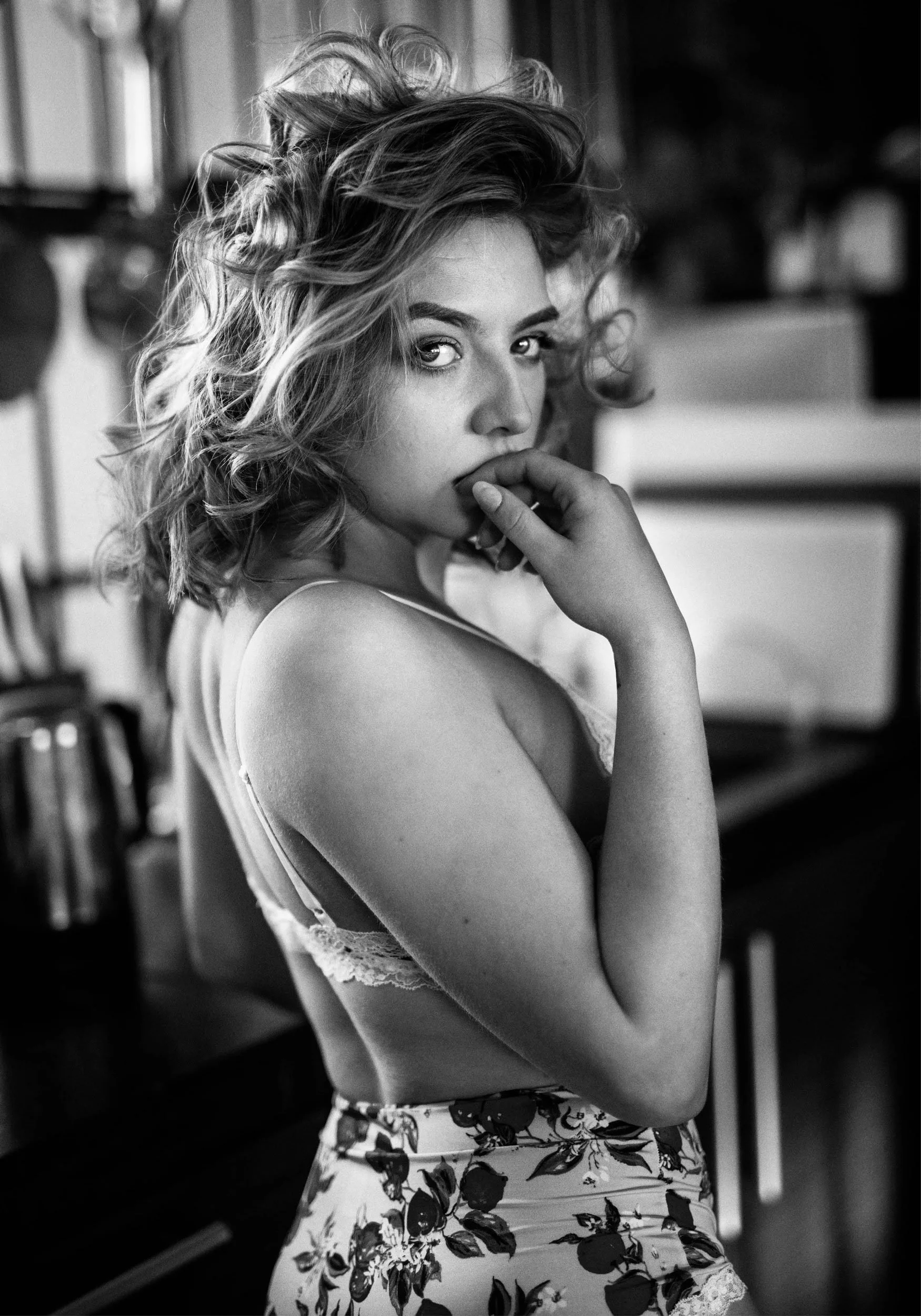 Black and white photo of a young woman with curly hair, wearing a lace top and floral skirt, looking over her shoulder while touching her lips with her fingers, in a room with kitchen appliances in the background.