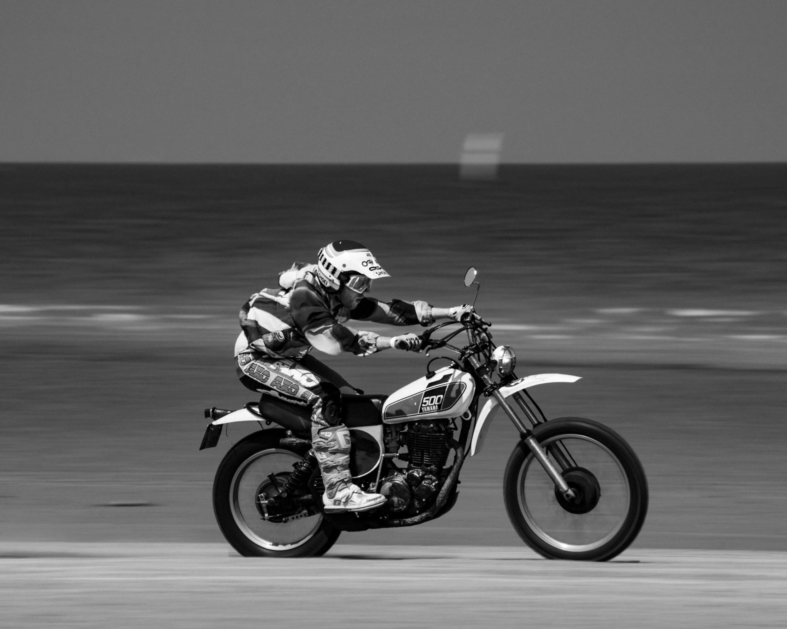 A person riding a Yamaha dirt bike on a road, with a helmet and protective gear, in black and white.