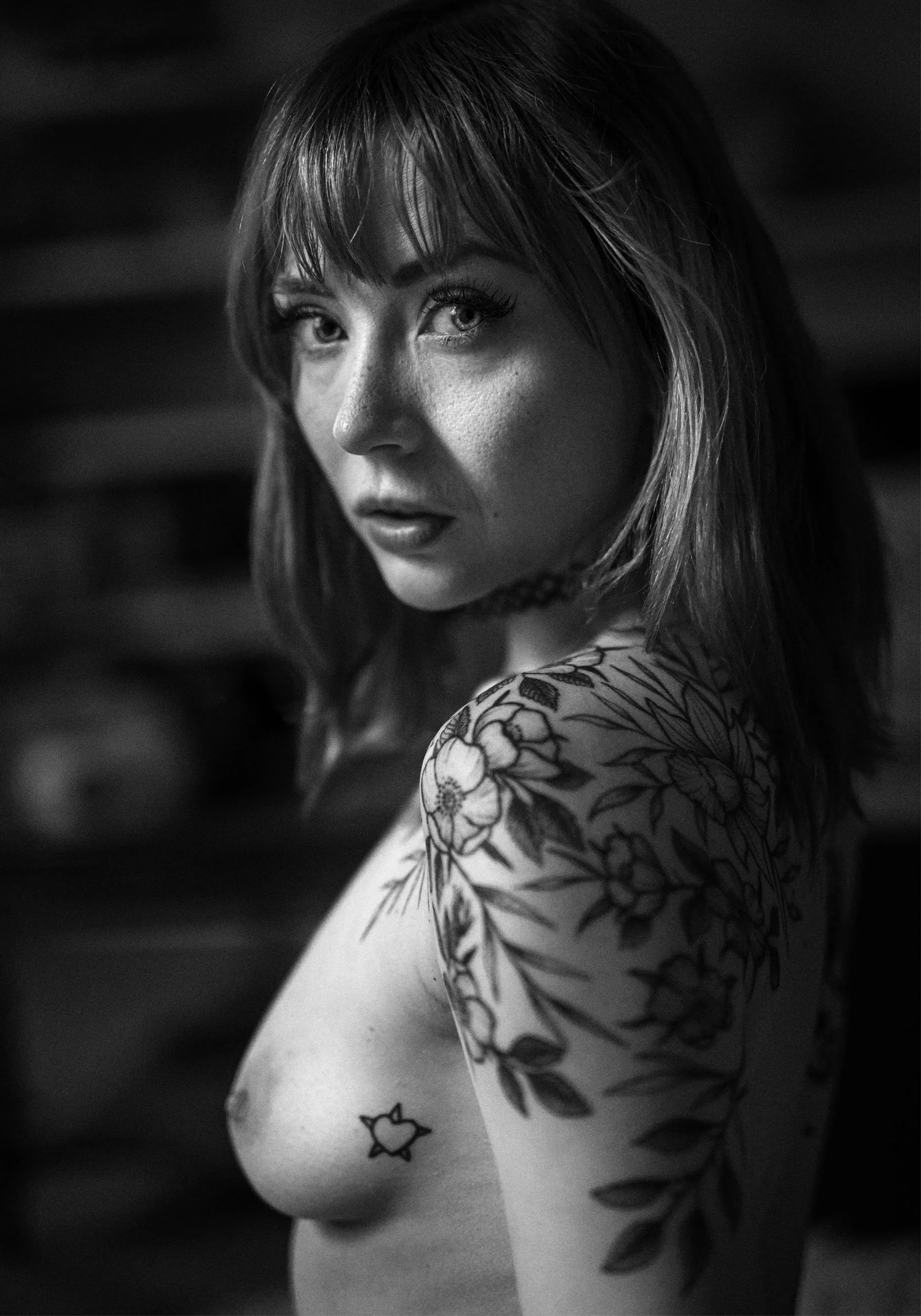 Black and white photo of a woman with shoulder-length hair, tattoos on her arm and shoulder, wearing a choker, looking over her shoulder at the camera.