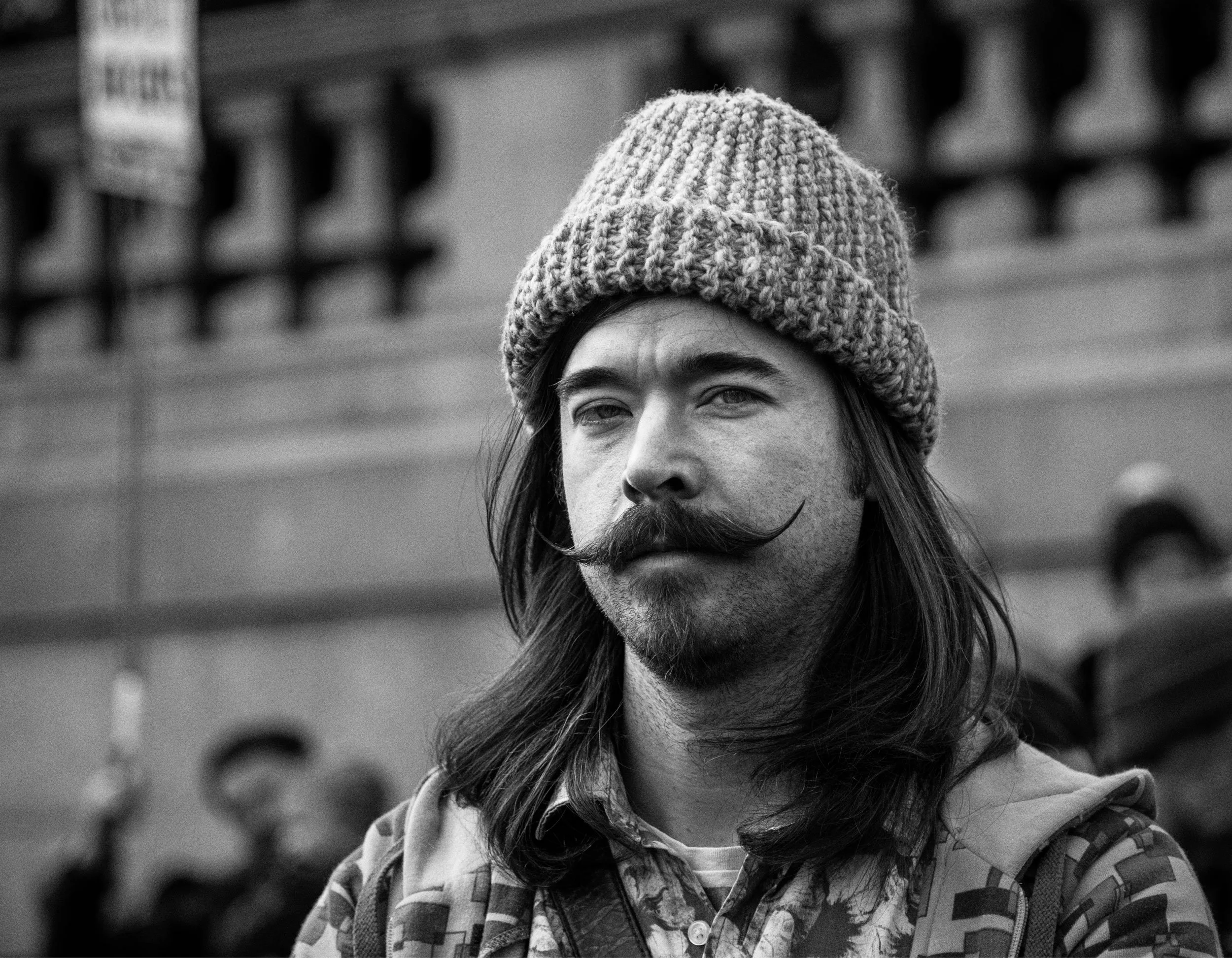 A black-and-white photograph of a man with long hair, a curled mustache, and a beard, wearing a knit beanie and patterned shirt, standing outdoors.