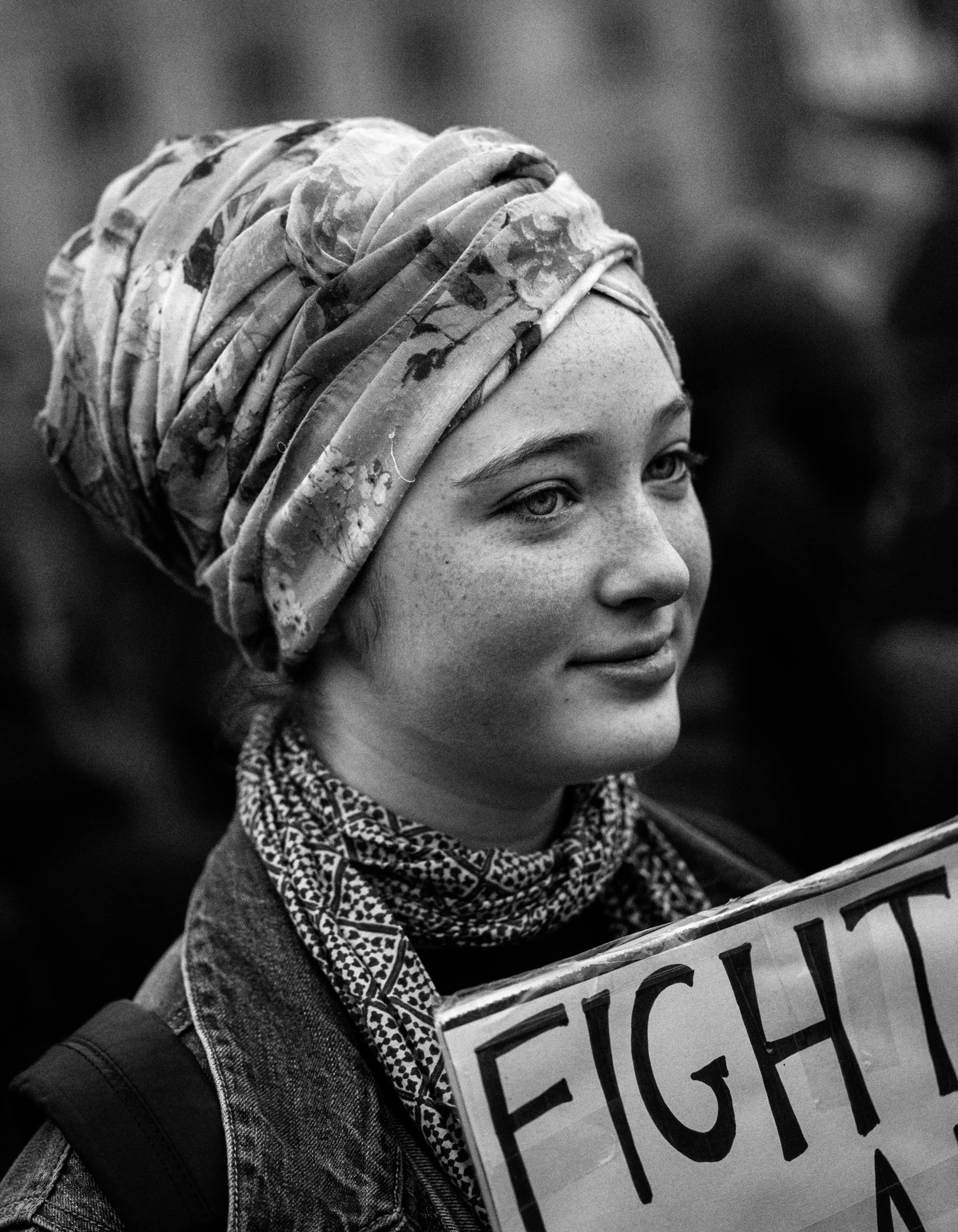 A young woman with light skin and freckles wearing a headscarf and patterned scarf, holding a protest sign with partially visible large text.