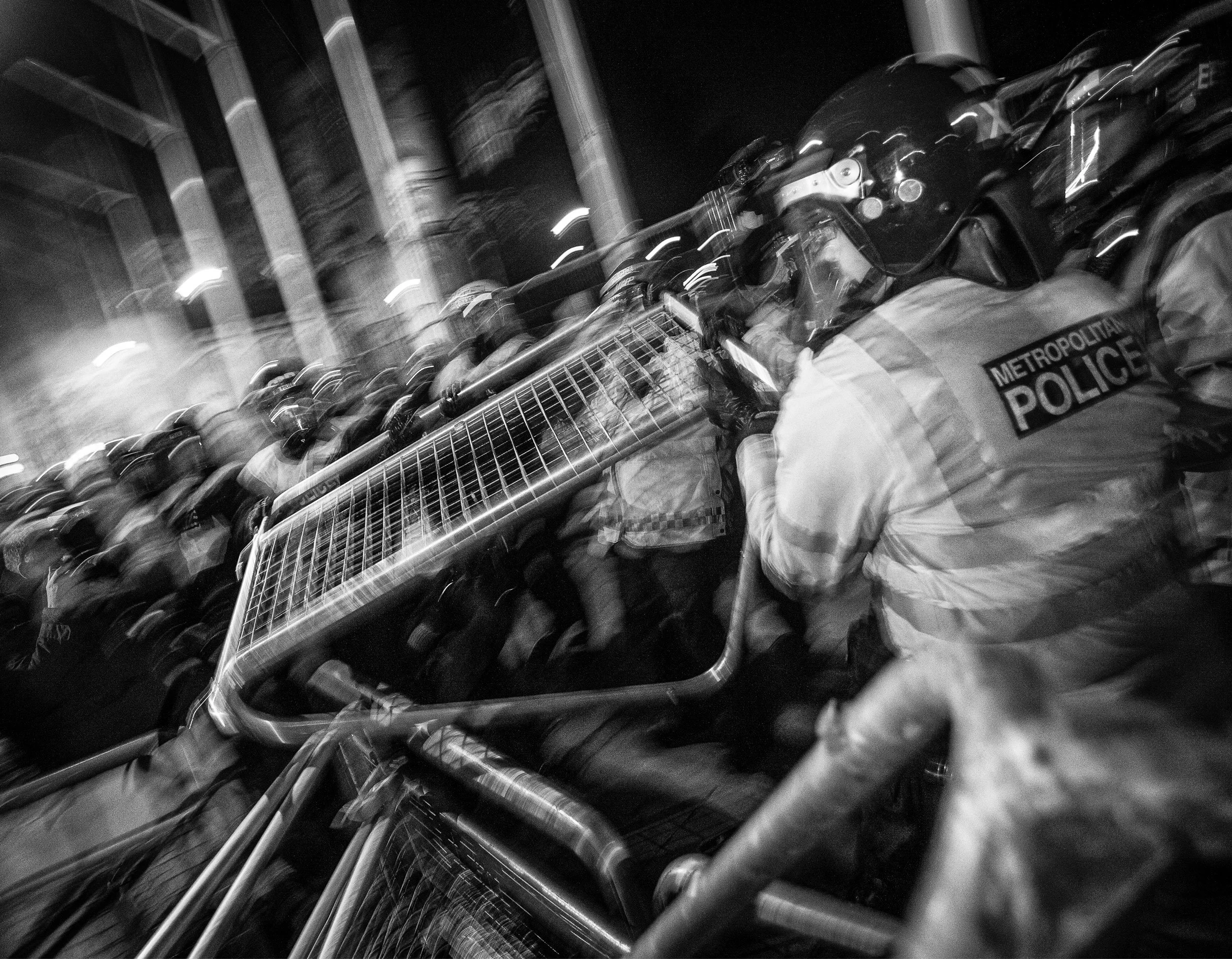 Nighttime scene of police officers in riot gear behind metal barricades, with some officers holding shields, part of the Metropolitan Police, in an urban area.