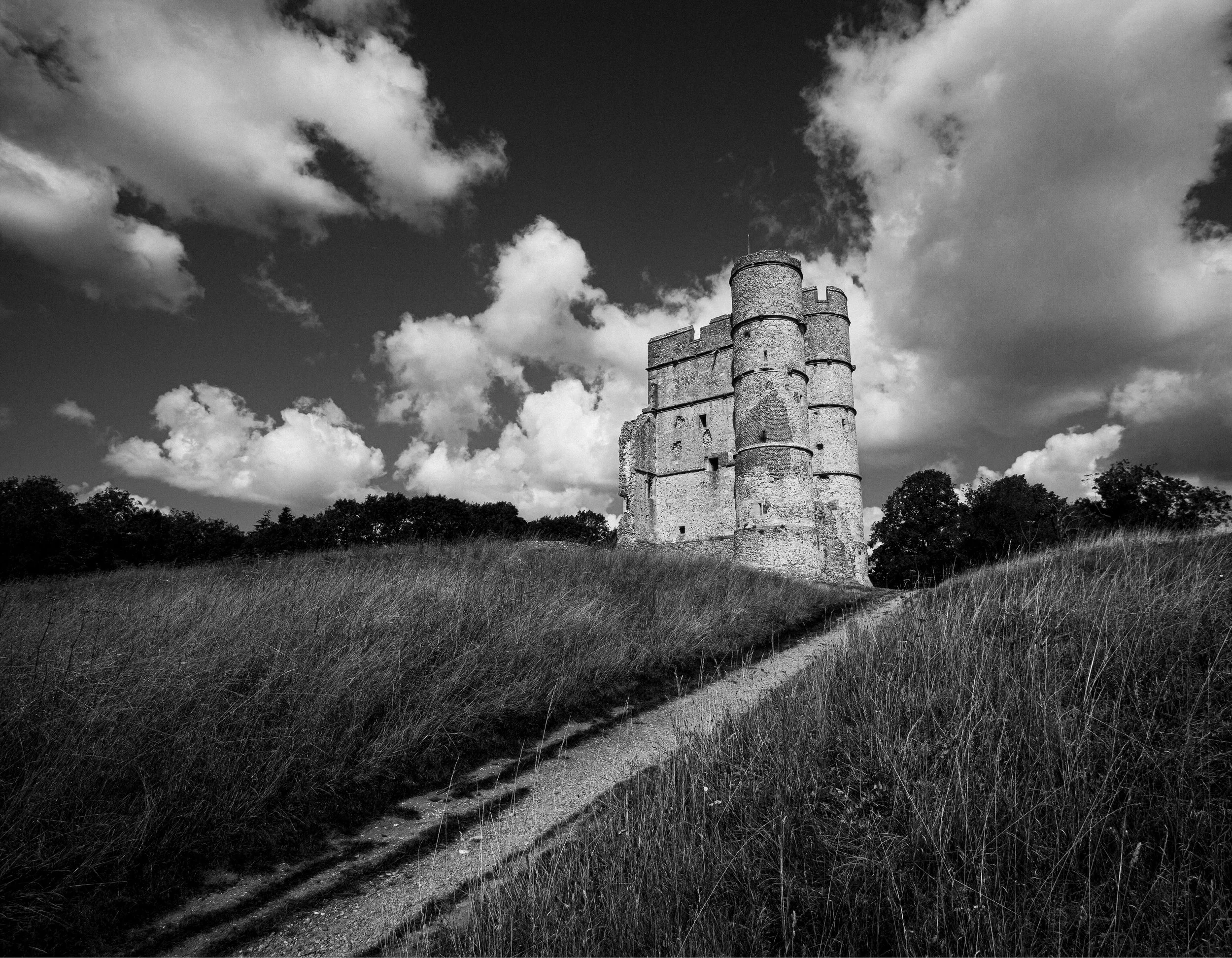 Black and white photo of a medieval castle tower on a grassy hill with a dirt path leading up to it, under a cloudy sky.