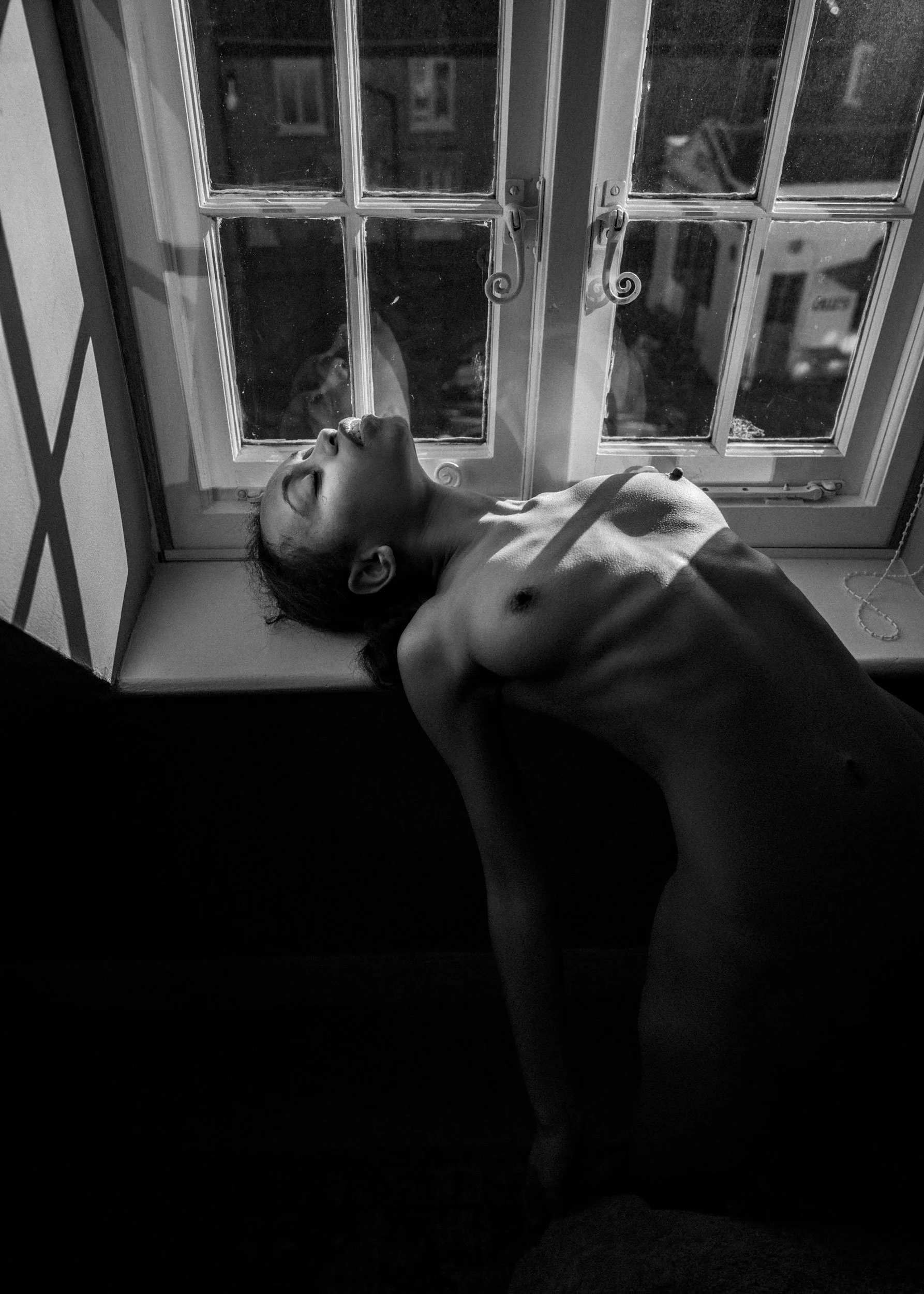 A black and white photo of a topless woman with closed eyes, lying on a windowsill with sunlight casting shadows on her face and body, in front of a window with panes and a latch.