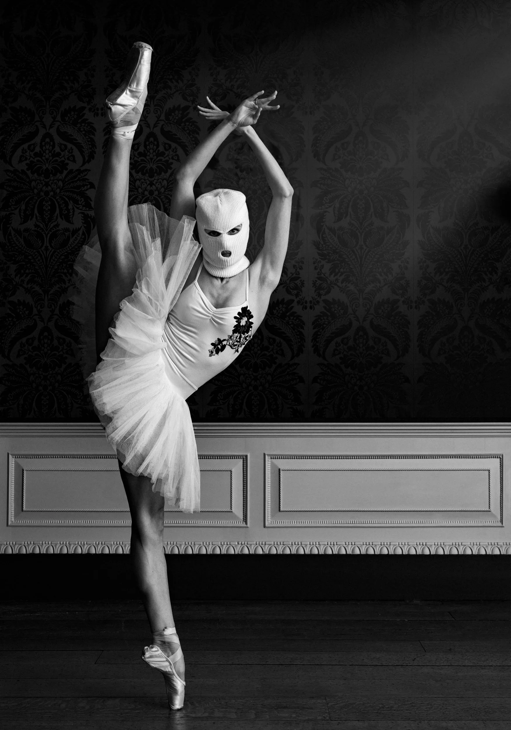 A dancer in a tutu and ballet shoes performs a standing split, wearing a ski mask, against a dark ornate wallpaper.